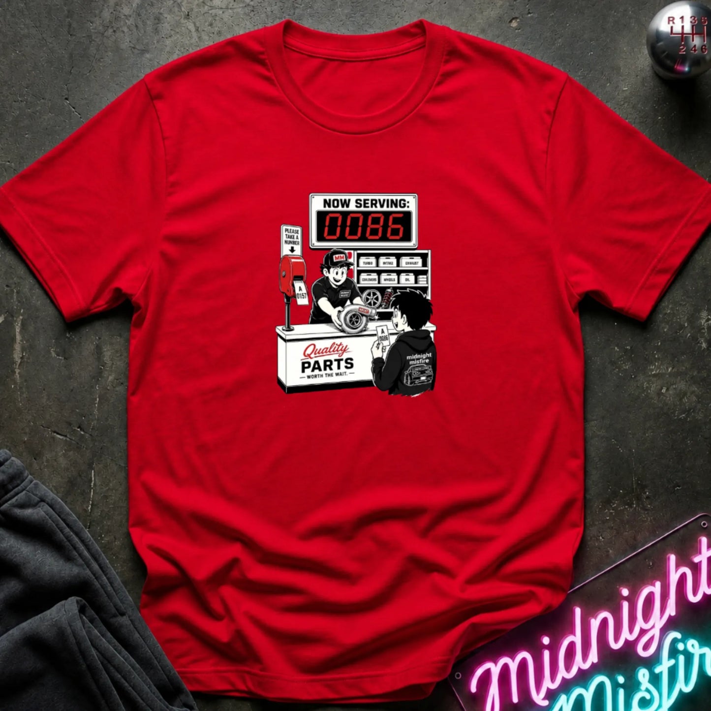 A red  short-sleeve t-shirt with a retro-style graphic of a garage attendant at a 'Quality Parts' counter, displayed flat on a dark textured surface with a neon sign and sweatpants partially visible.