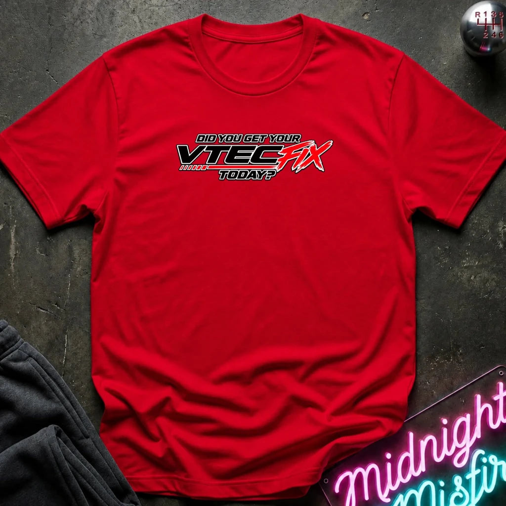 A red short-sleeve crewneck t-shirt with a graphic print reading 'DID YOU GET YOUR VTEC FIX TODAY?' in black, white, and red. The shirt is laid flat on a dark textured surface with a gray sweatshirt corner and a neon sign visible as props.