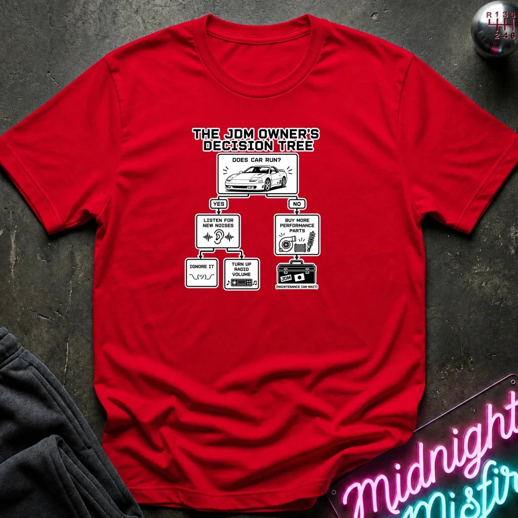 A red short-sleeve graphic t-shirt laid flat on a dark textured surface. The shirt features a white and black printed graphic titled 'THE JDM OWNER'S DECISION TREE' with flowchart boxes and illustrations related to car maintenance and modifications.