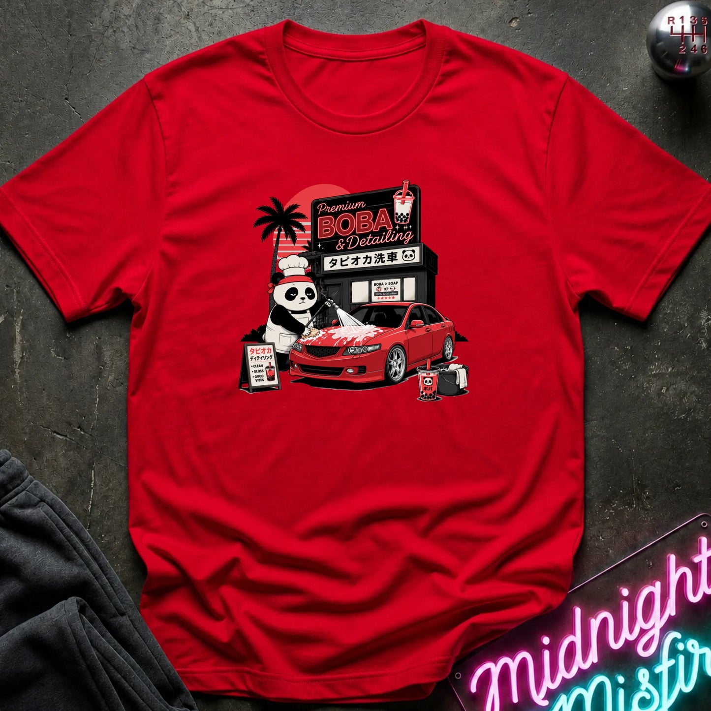 A red  short-sleeve graphic T-shirt laid flat on a dark textured surface. The front print shows a red car, a panda character, and signage reading 'Premium DOBA Detailing' with Japanese text.