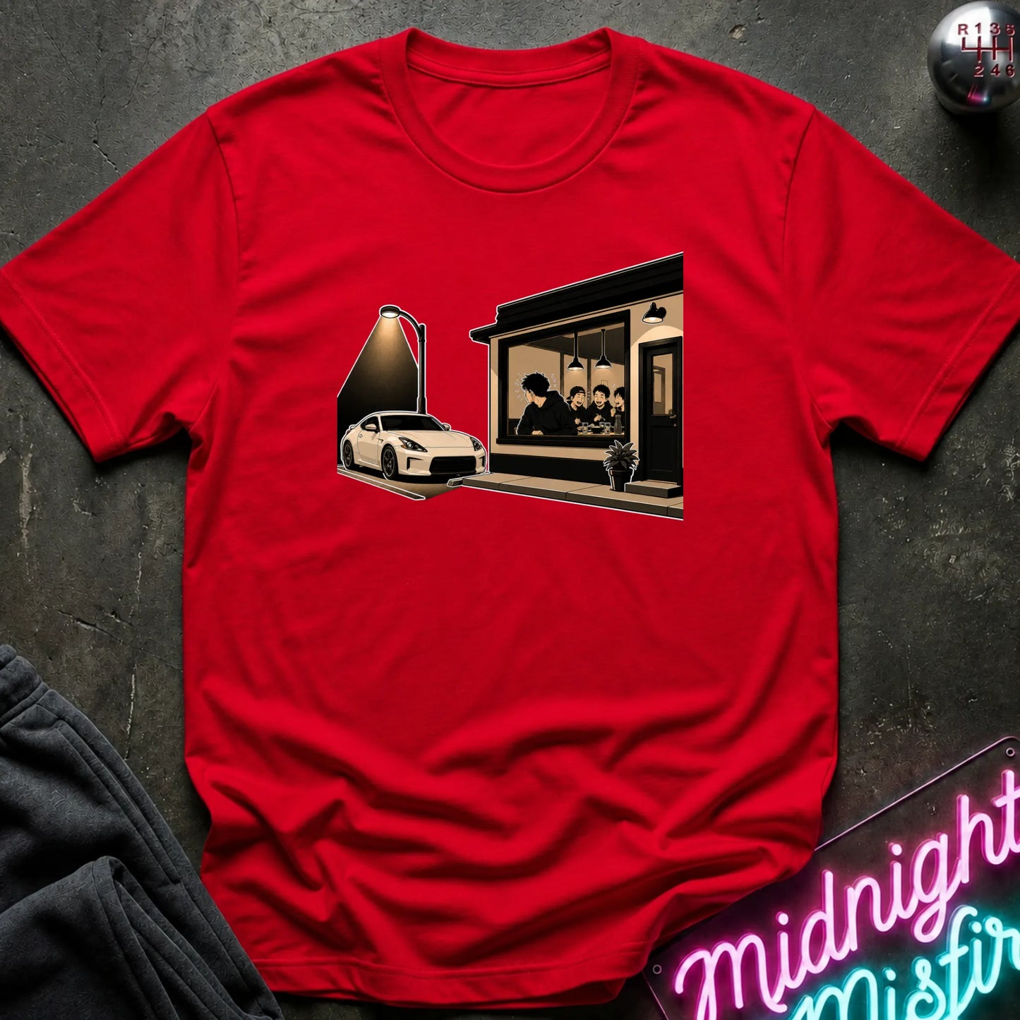 A red short-sleeve crewneck t-shirt laid flat on a dark textured surface. The shirt features a graphic print in the center showing a white sports car under a spotlight next to a storefront with silhouetted figures.