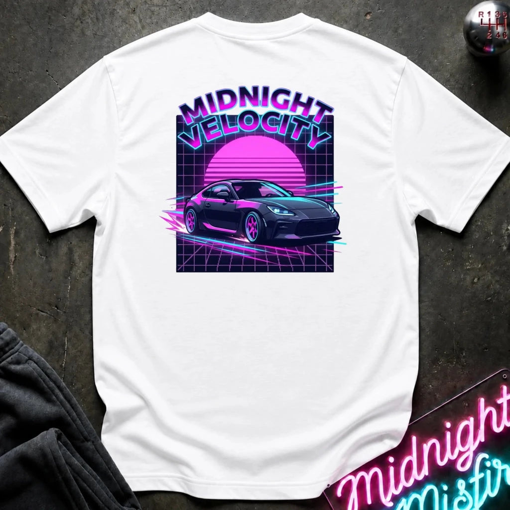 A white short-sleeve t-shirt laid flat on a dark textured surface, featuring a retro 80s synthwave graphic on the back with the text 'MIDNIGHT VELOCITY', a pink sun, and a dark sports car. A neon sign and dark pants are partially visible as props.