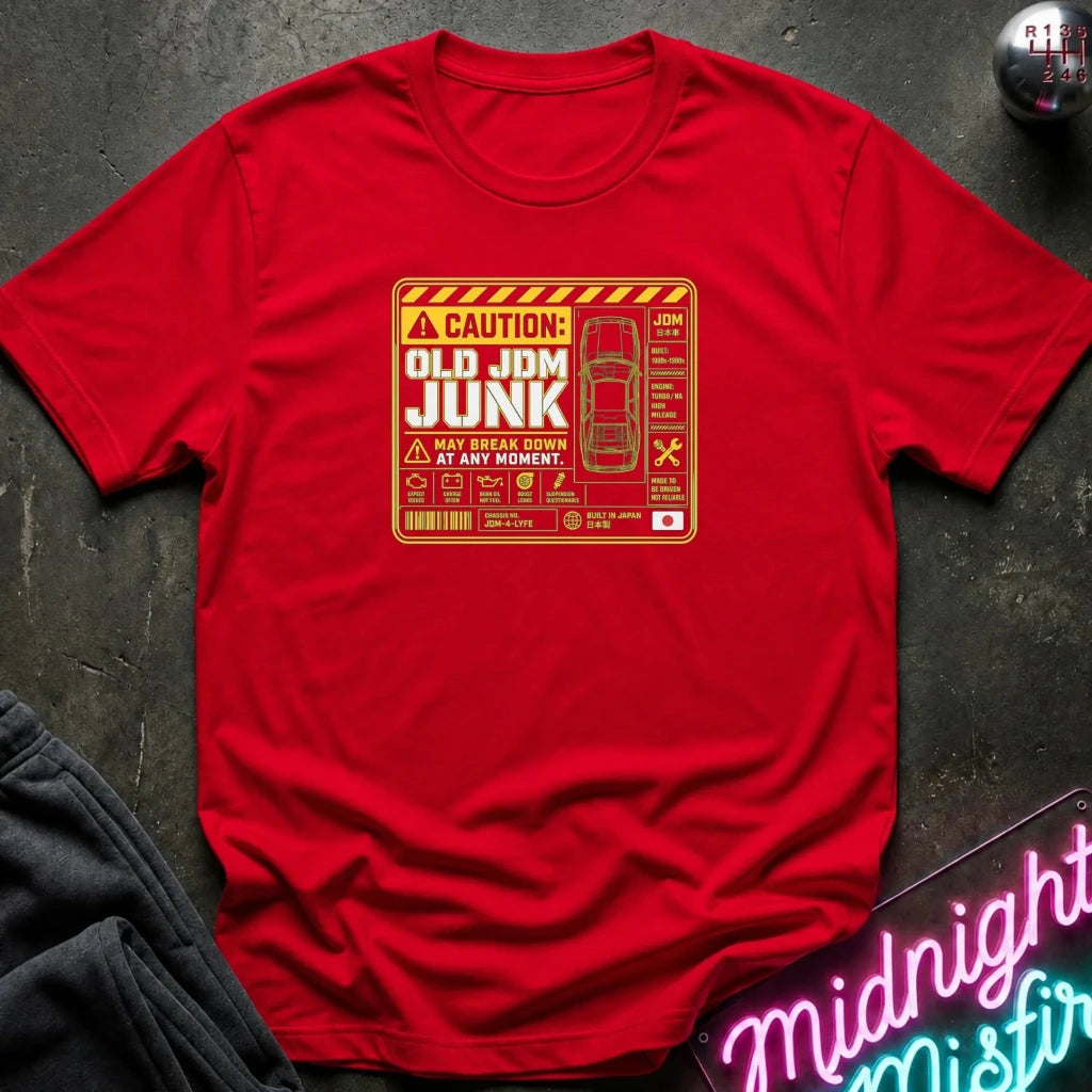 A red short-sleeve crewneck t-shirt laid flat on a dark textured surface, featuring a retro-style orange and yellow caution sign graphic on the chest that reads 'OLD JDM JUNK'.