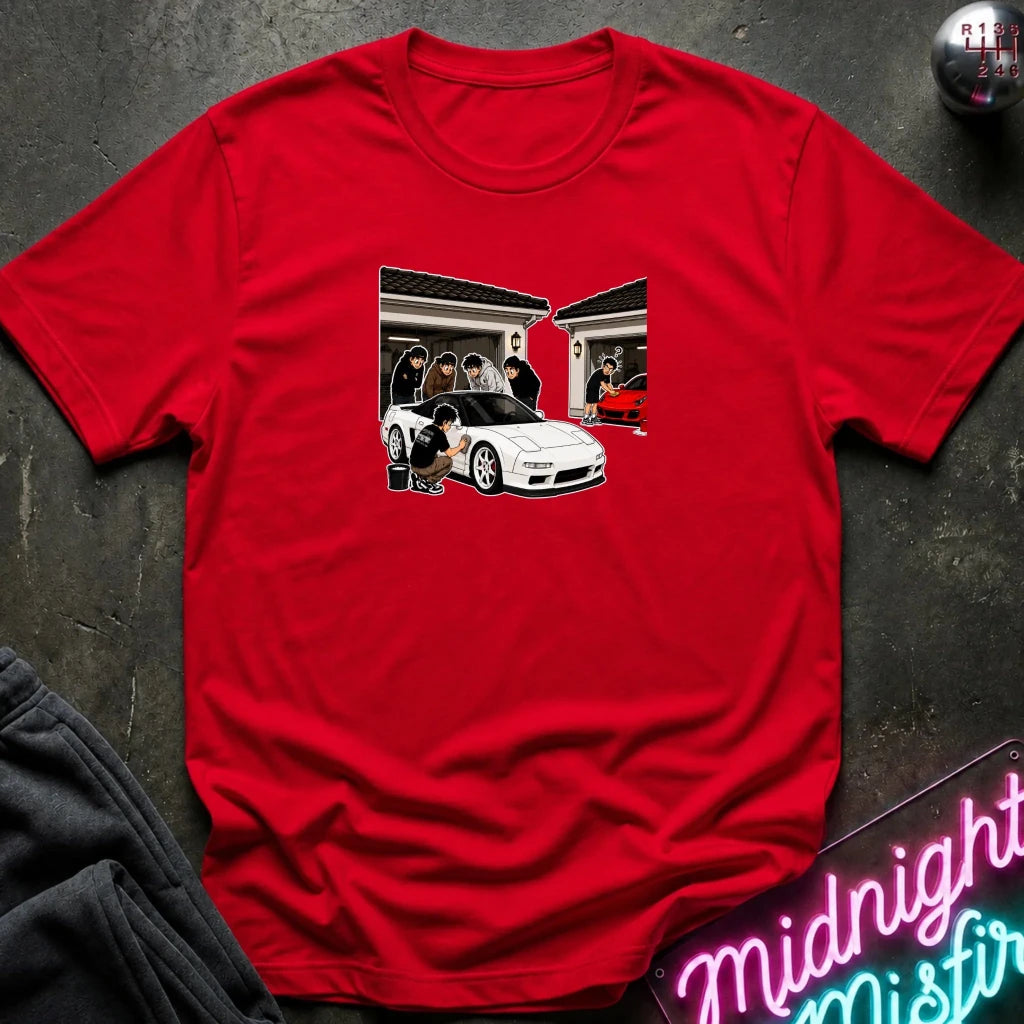 A red short-sleeve crewneck t-shirt laid flat on a dark textured surface, featuring a graphic print of a white sports car in a garage scene.
