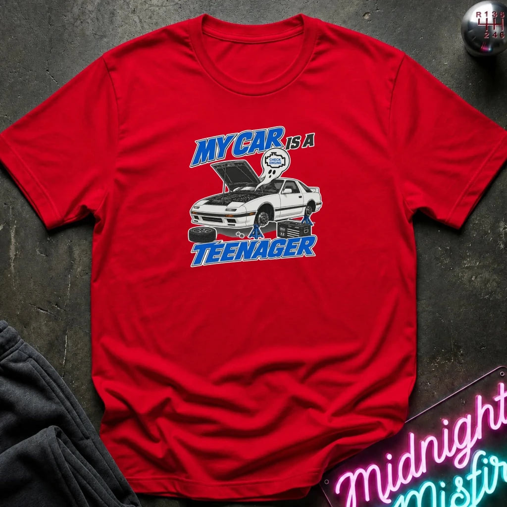 A red short-sleeve graphic T-shirt laid flat on a dark textured surface. The shirt features a white car illustration with its hood open, surrounded by blue and white text that reads 'MY CAR IS A TEENAGER.