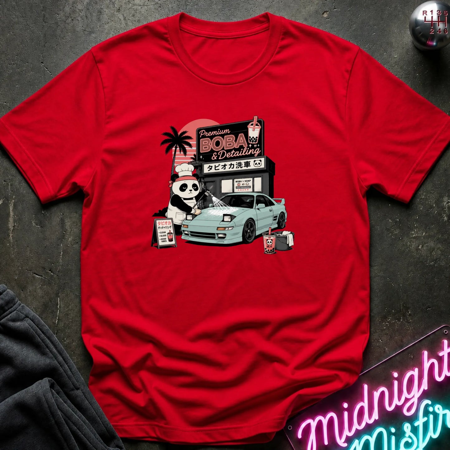 A red short-sleeve graphic T-shirt laid flat on a dark textured surface. The shirt features a colorful retro-style print of a panda character, a light blue sports car, and a sign reading 'Premium BOBA & Coffee' with Japanese text.