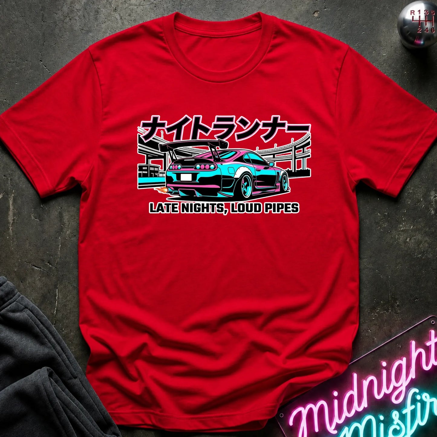 A red short-sleeve graphic T-shirt laid flat on a dark textured surface. The shirt features a central graphic of a teal and black sports car with Japanese text above and the phrase 'LATE NIGHTS, LOUD PIPES' below.