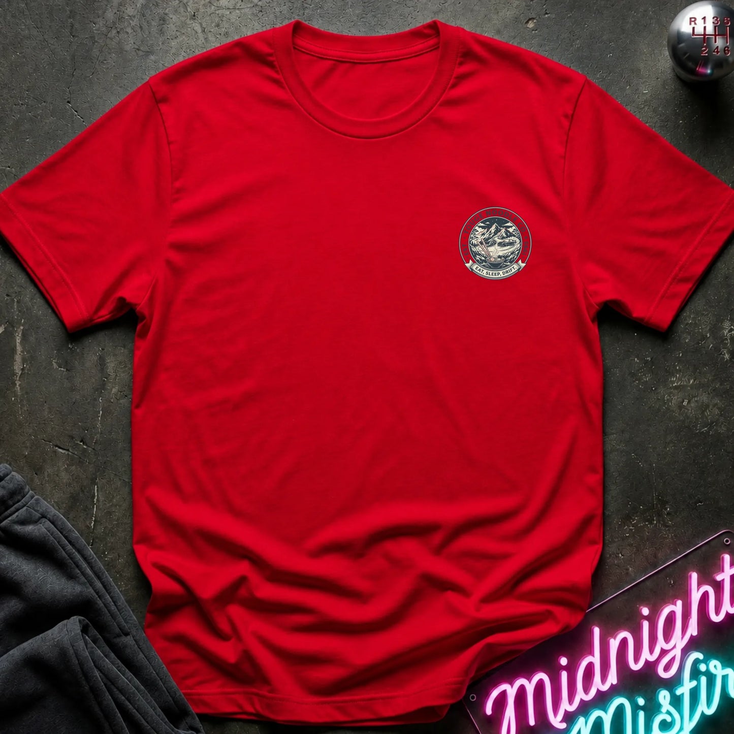 Red crewneck t-shirt with a circular crest printed on the left chest, styled flat on a dark textured surface with a neon sign and dark pants in the background.