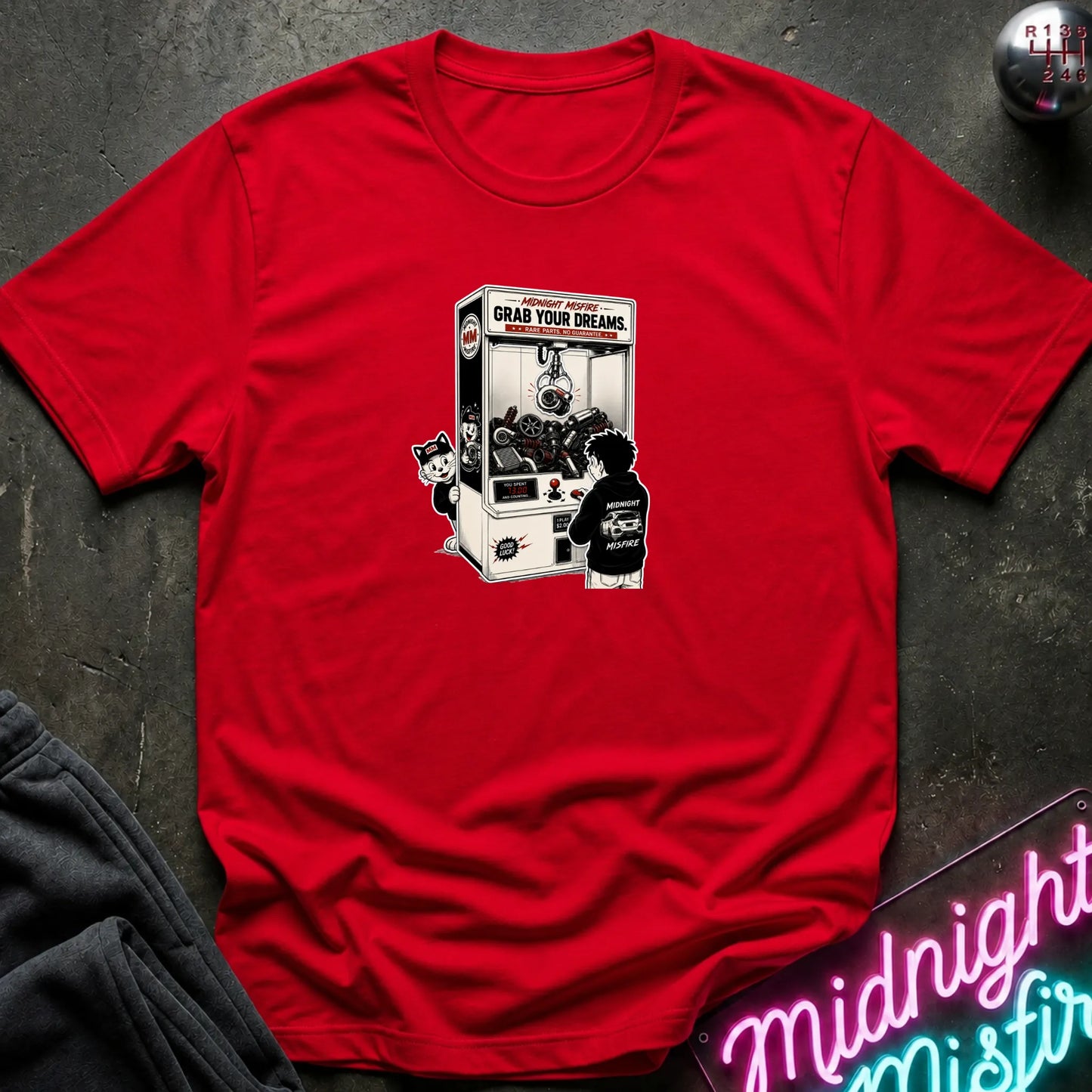 A red short-sleeve crewneck t-shirt laid flat on a dark textured surface, featuring a central graphic of a person in a black hoodie playing a claw machine. 