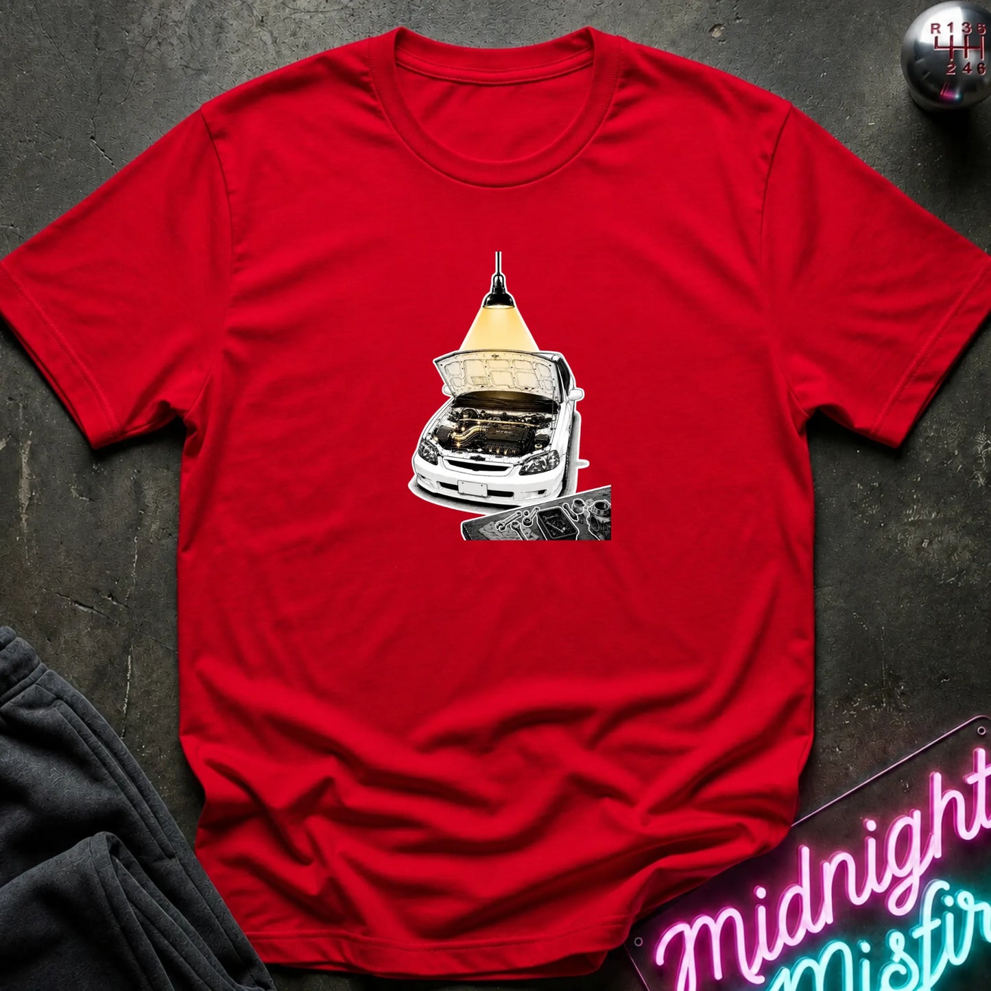 A red short-sleeve t-shirt laid flat on a dark textured surface, featuring a graphic print of a white car with its hood open under a glowing yellow lamp. 