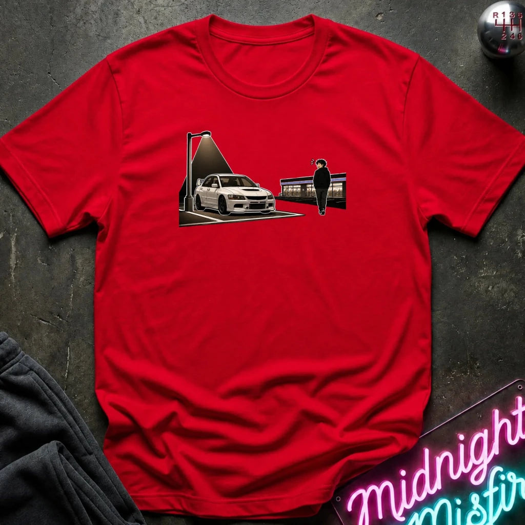 A red short-sleeve crewneck t-shirt laid flat on a dark textured surface, featuring a graphic print of a white sports car under a streetlamp next to a figure. Styled flat-lay props include a gray sweatshirt corner, a pink neon sign, and a gear shift knob.
