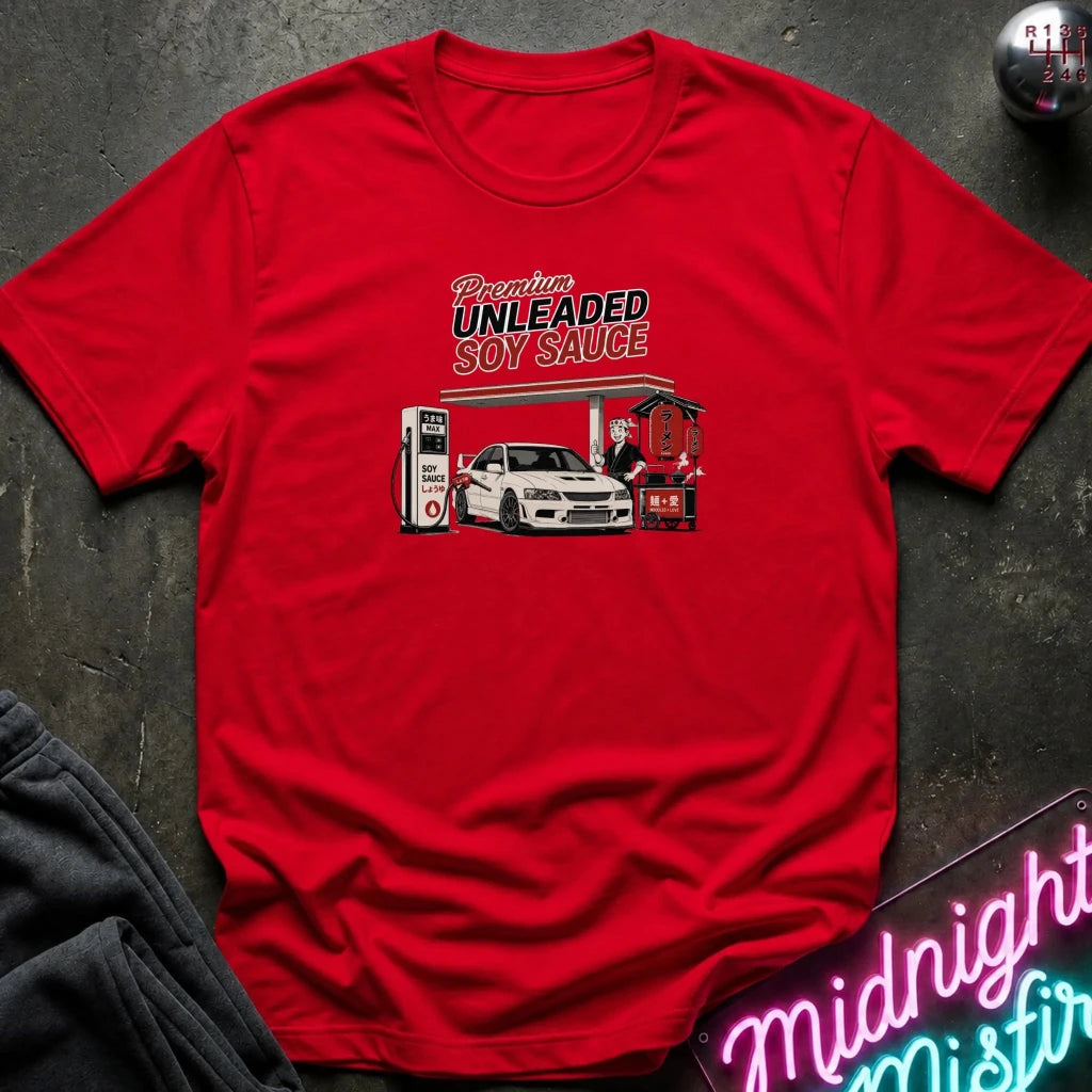 A red short-sleeve graphic T-shirt laid flat on a dark textured surface. The shirt features a vintage-style illustration of a white sports car at a gas station with the text 'Premium UNLEADED SOY SAUCE.