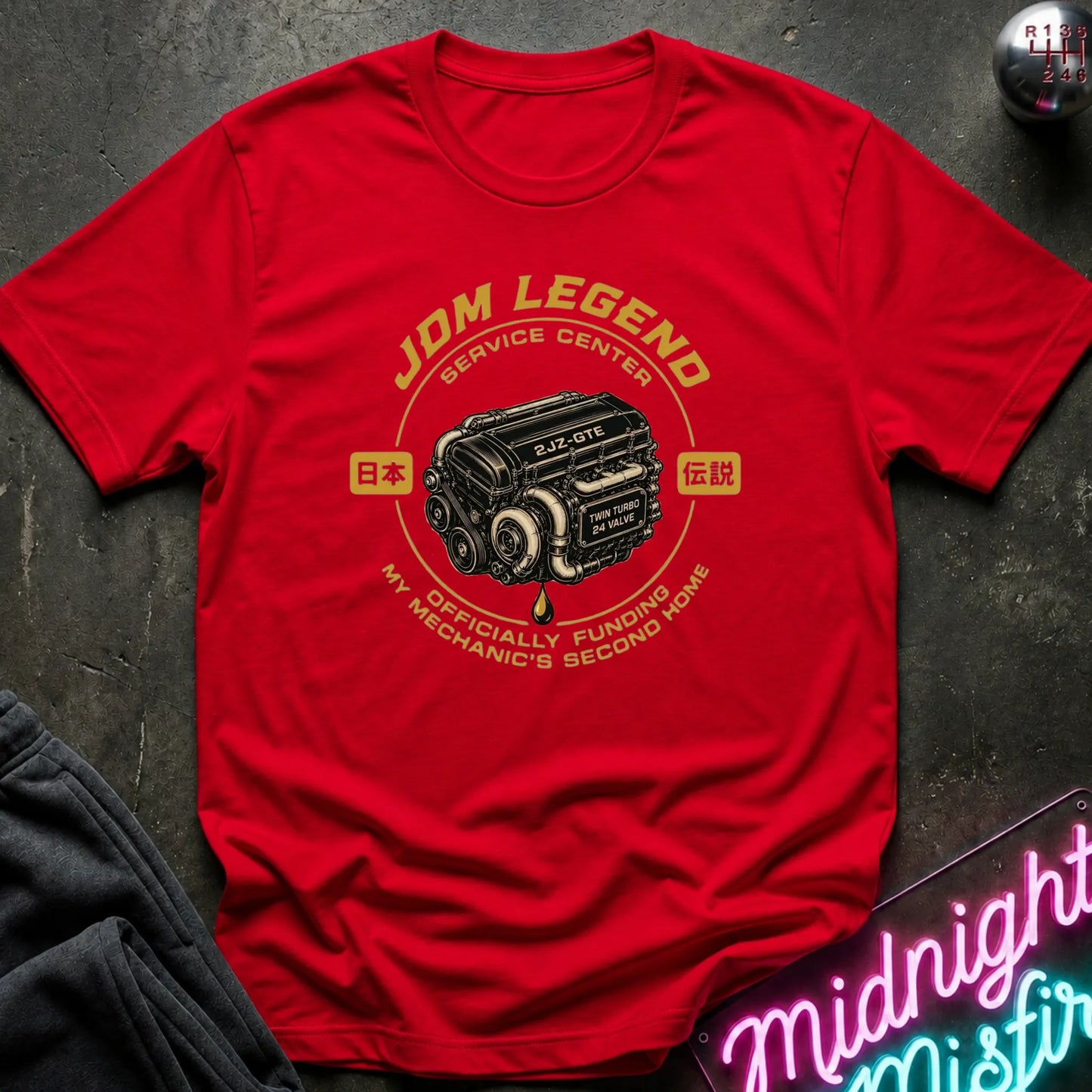 A red short-sleeve graphic T-shirt laid flat on a dark textured surface. The shirt features a vintage-style chest print with the words 'JDM LEGEND SERVICE CENTER' and a black engine illustration, plus smaller text reading 'MY MECHANIC'S SECOND HOME.