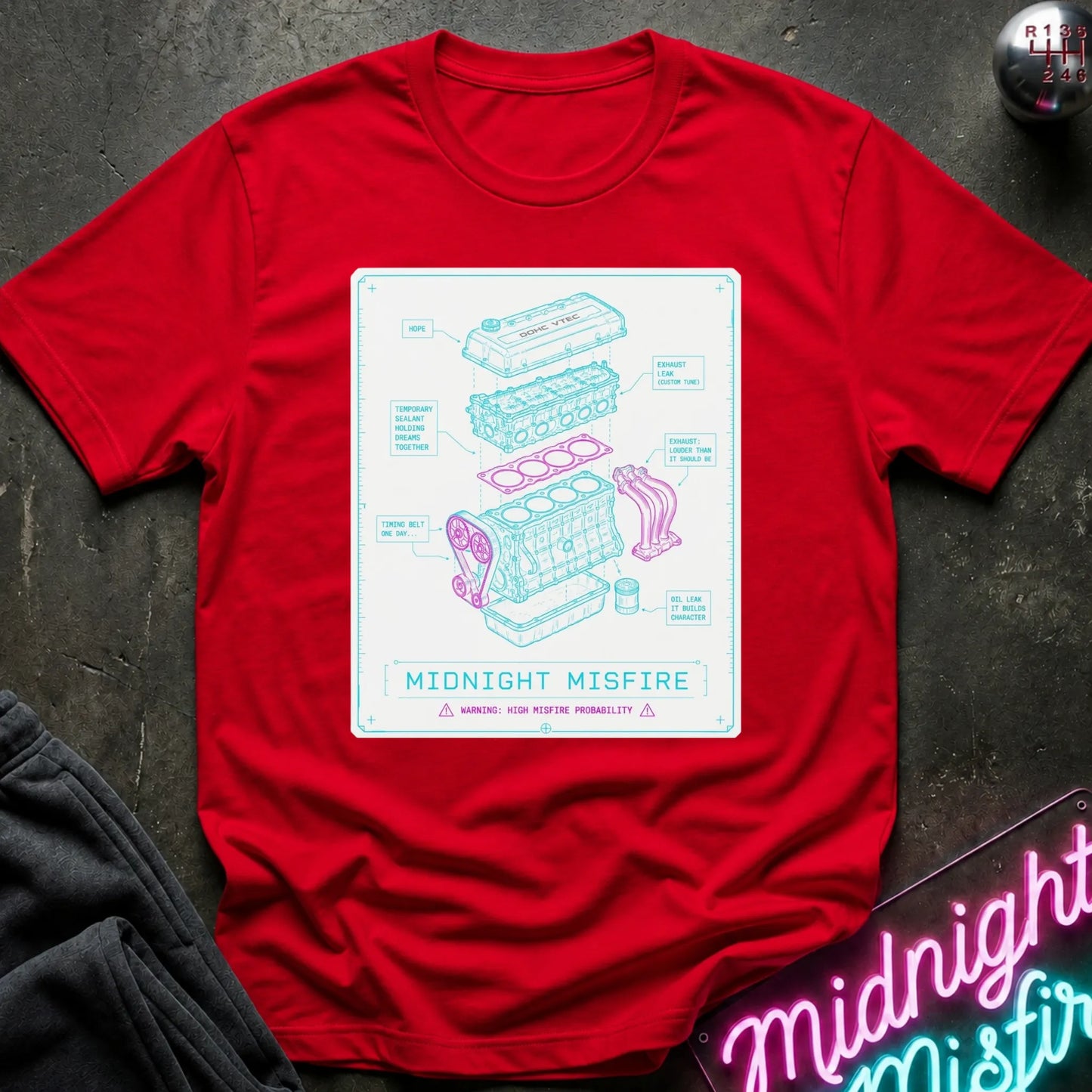 A red short-sleeve crewneck t-shirt laid flat on a dark textured surface. The center of the shirt features a white rectangular graphic with a light blue and pink exploded-style technical illustration and the text 'MIDNIGHT MISFIRE'.