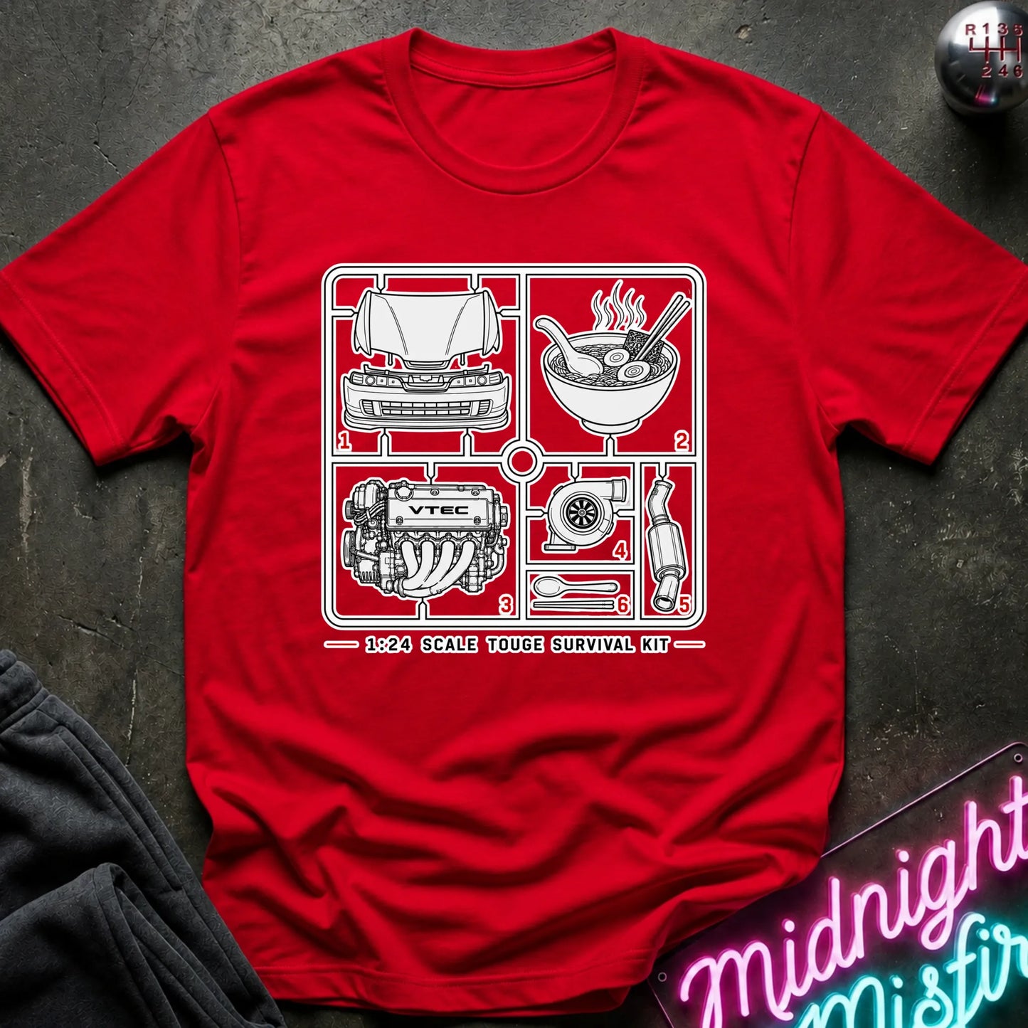 A red short-sleeve graphic T-shirt laid flat on a dark textured surface. The shirt features a black line-art schematic of a survival kit with numbered illustrations and the text '1:24 SCALE TOUGHE SURVIVAL KIT' printed on the chest.