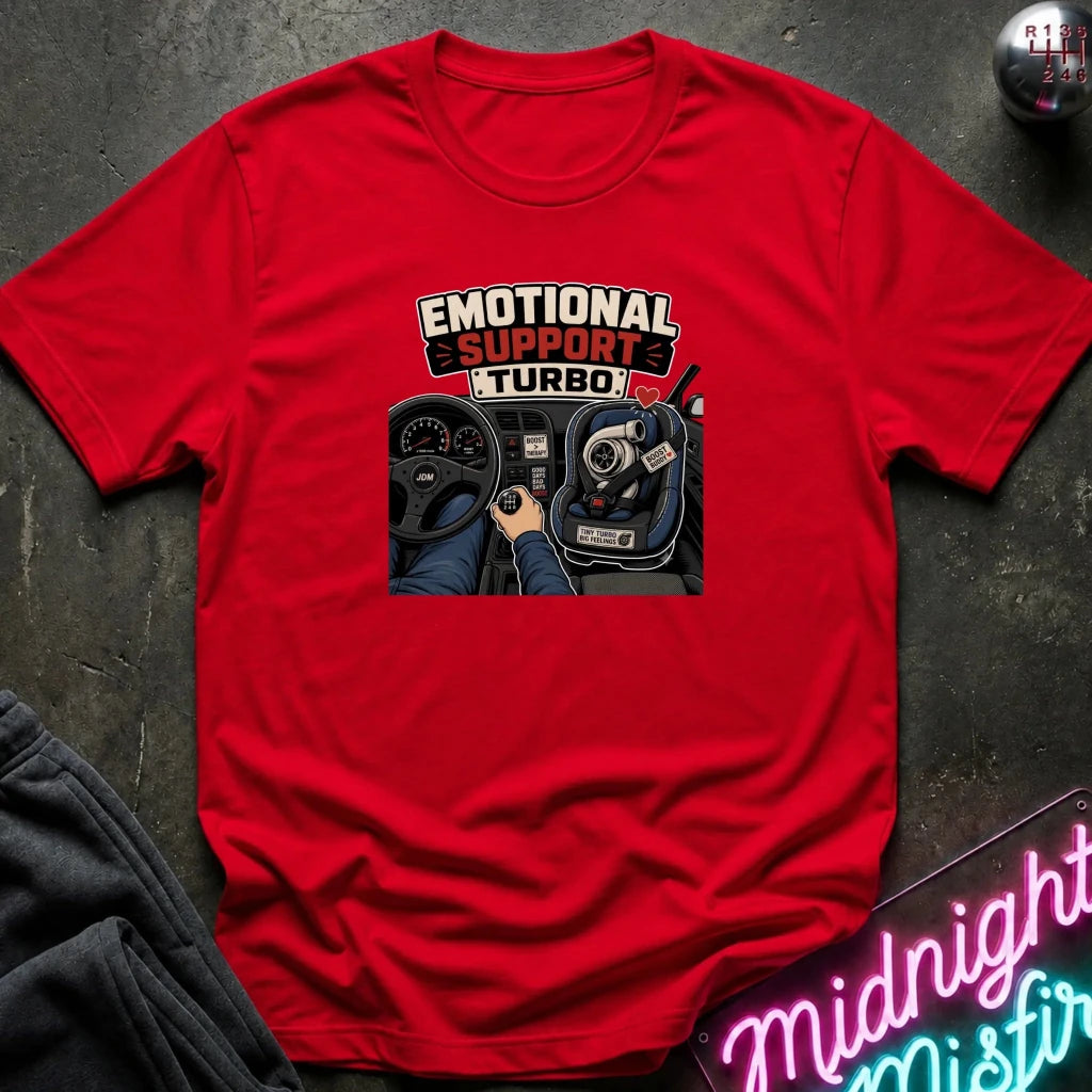 A red short-sleeve graphic T-shirt laid flat on a dark textured surface. The shirt features a central print with the words 'EMOTIONAL SUPPORT TURBO' above an illustration of a hand on a steering wheel and car dashboard.