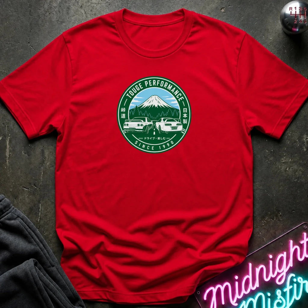 A red short-sleeve crewneck t-shirt laid flat on a dark textured surface. The center of the shirt features a circular green and white graphic depicting Mount Fuji