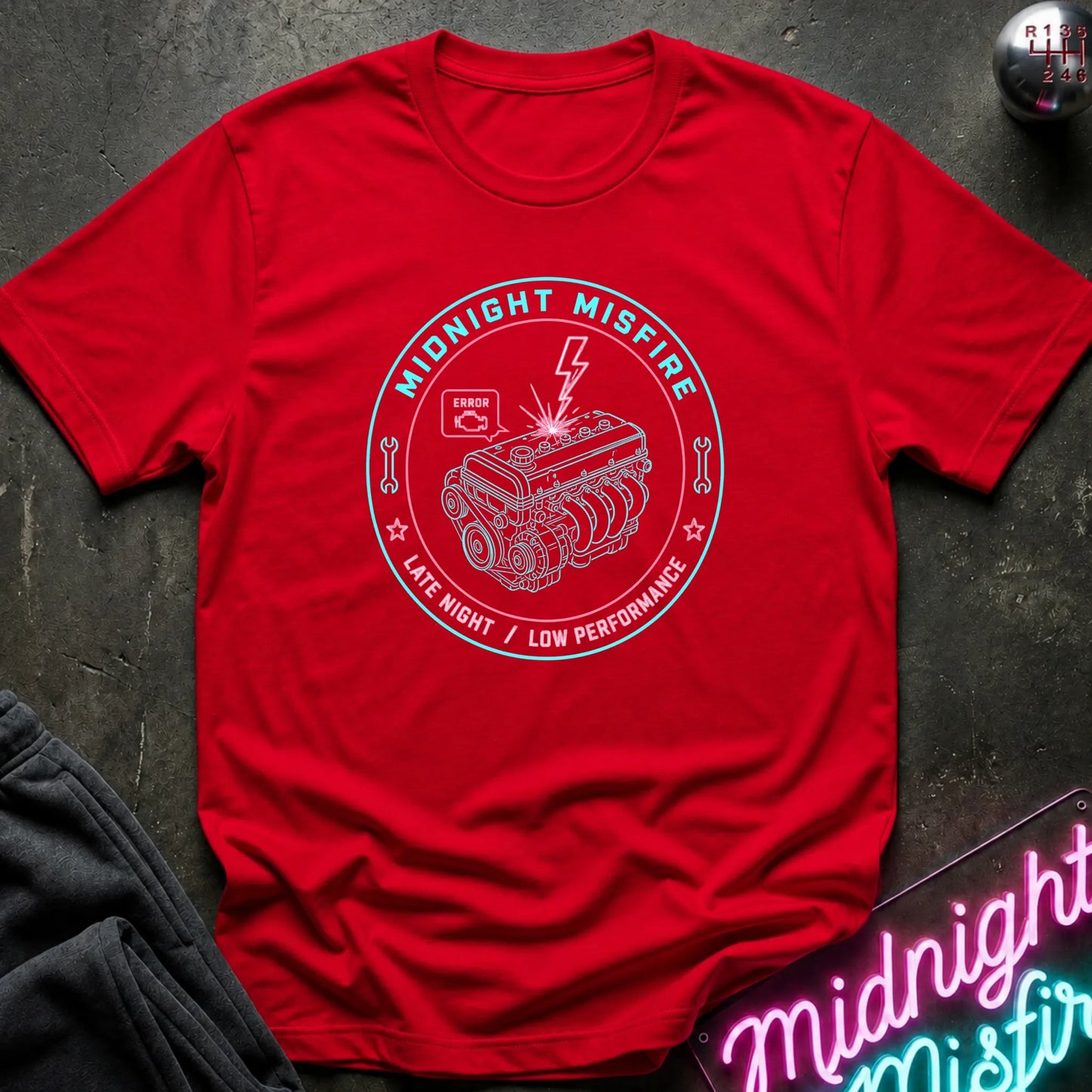 A red short-sleeve crewneck t-shirt with a white circular graphic on the chest depicting an engine and the text 'MIDNIGHT MISFIRE' and 'LATE NIGHT / LOW PERFORMANCE'. The shirt is laid flat on a dark textured surface with a neon sign and dark pants partially visible.