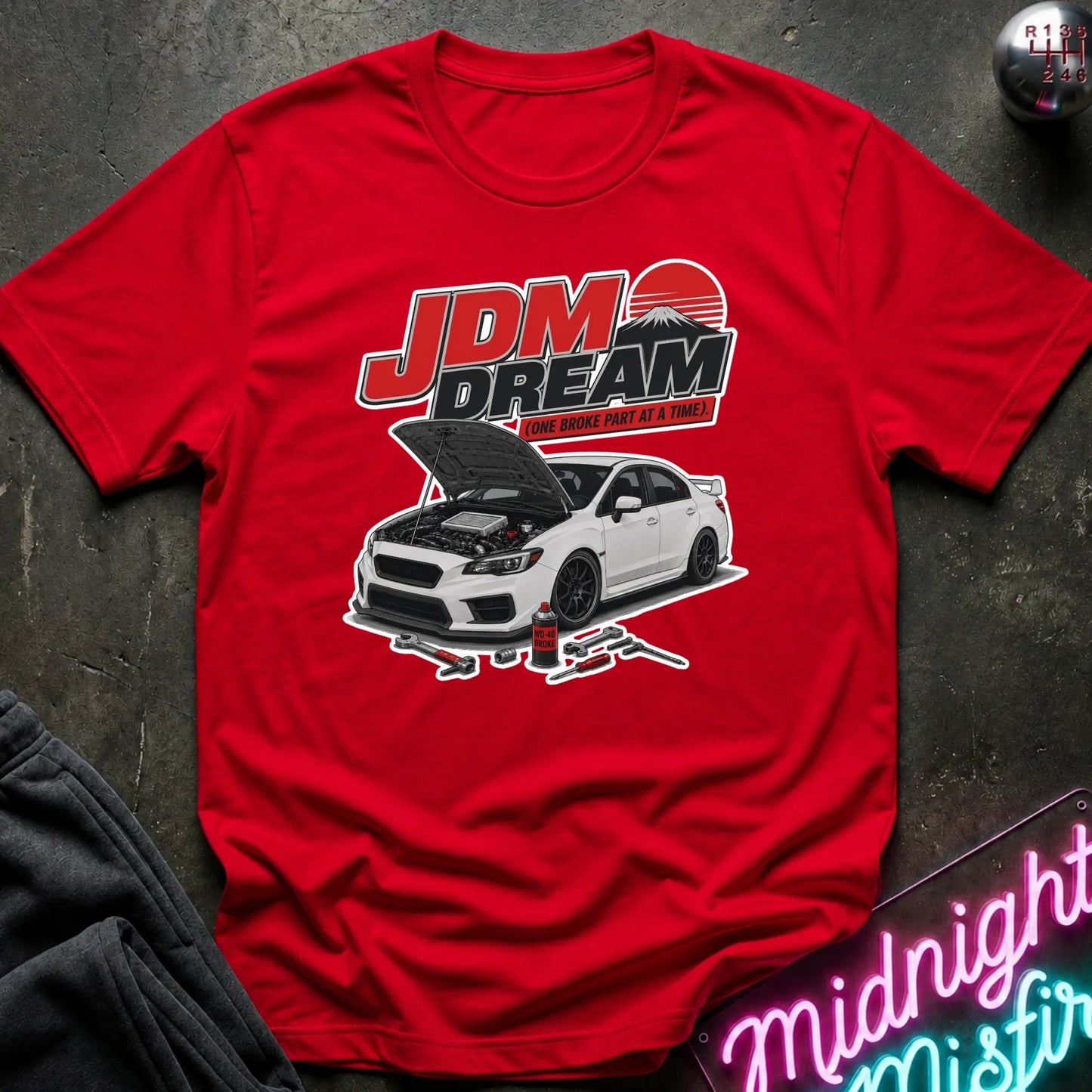 A red short-sleeve graphic T-shirt laid flat on a dark textured surface. The shirt features a large chest print with the text 'JDM DREAM' above a white sports car with its hood open, plus small tool graphics.