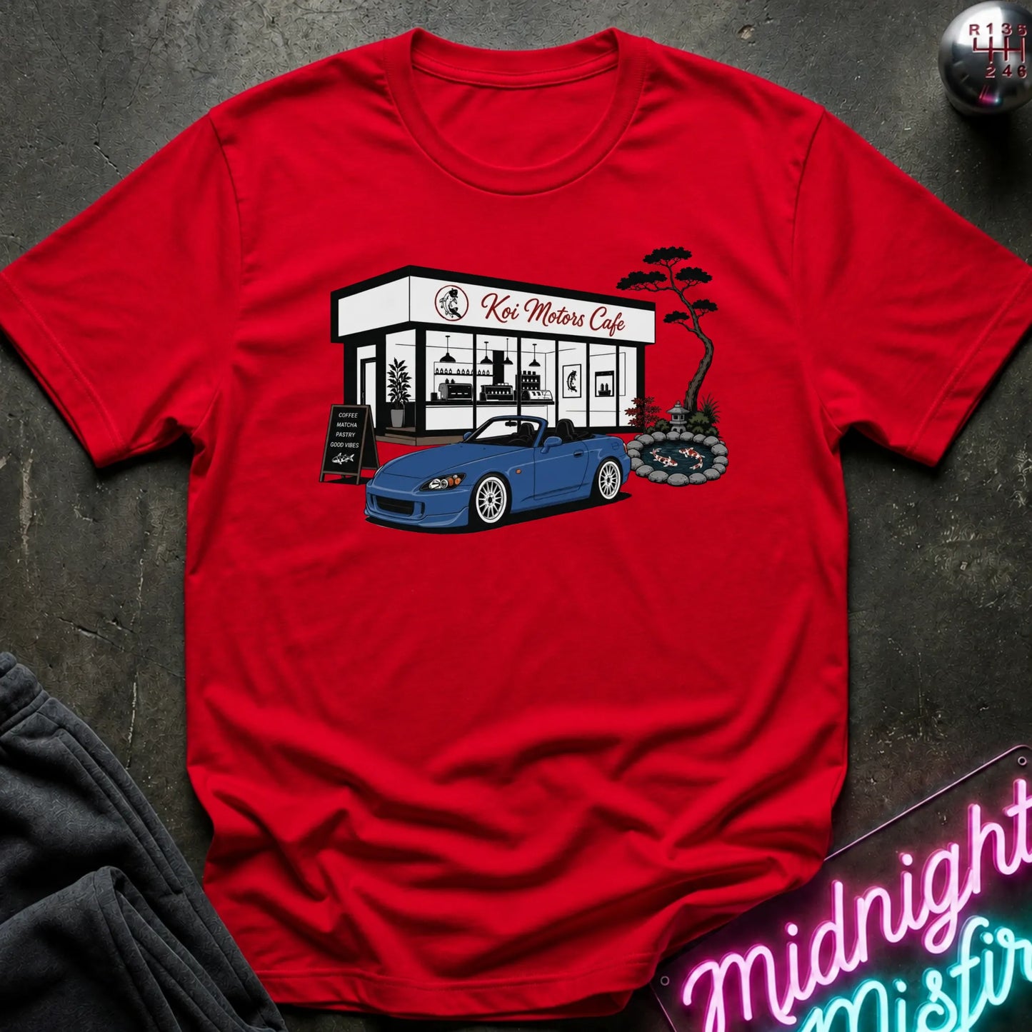A red short-sleeve crewneck t-shirt laid flat on a dark textured surface. The shirt features a graphic print of a blue convertible sports car parked in front of 'Koi Motor Cafe' with a small tree illustration.