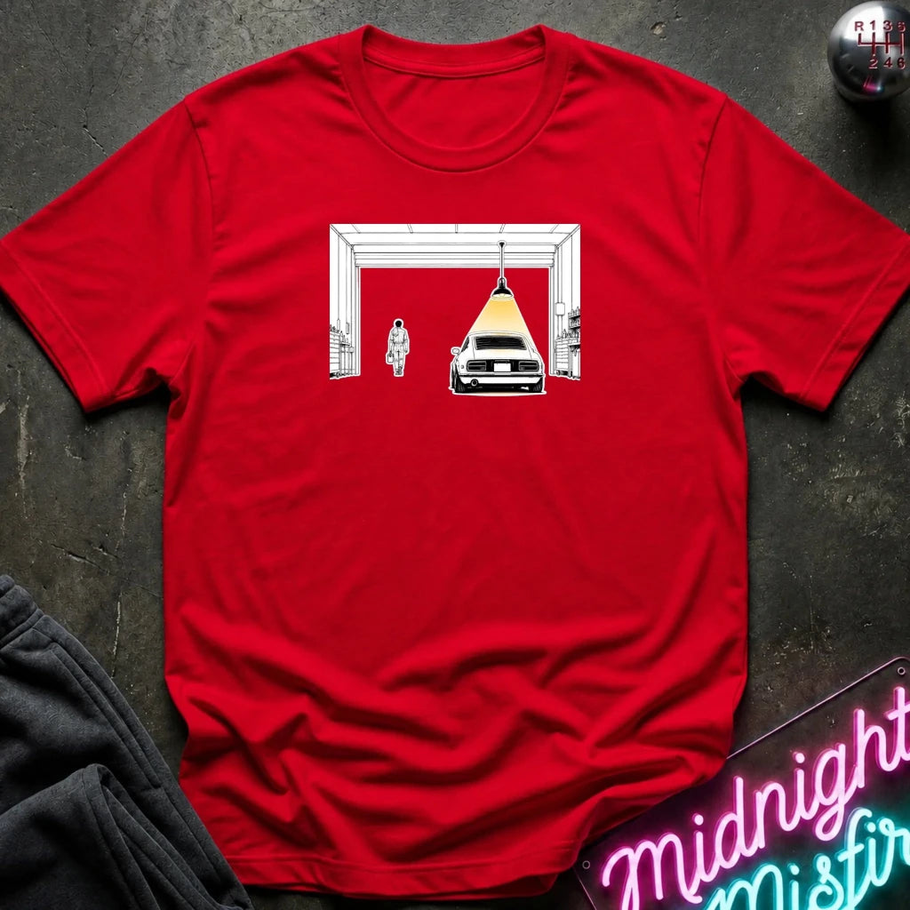 A red short-sleeve crewneck t-shirt laid flat on a dark textured surface, featuring a graphic print of a white car under a yellow spotlight inside a white frame. 