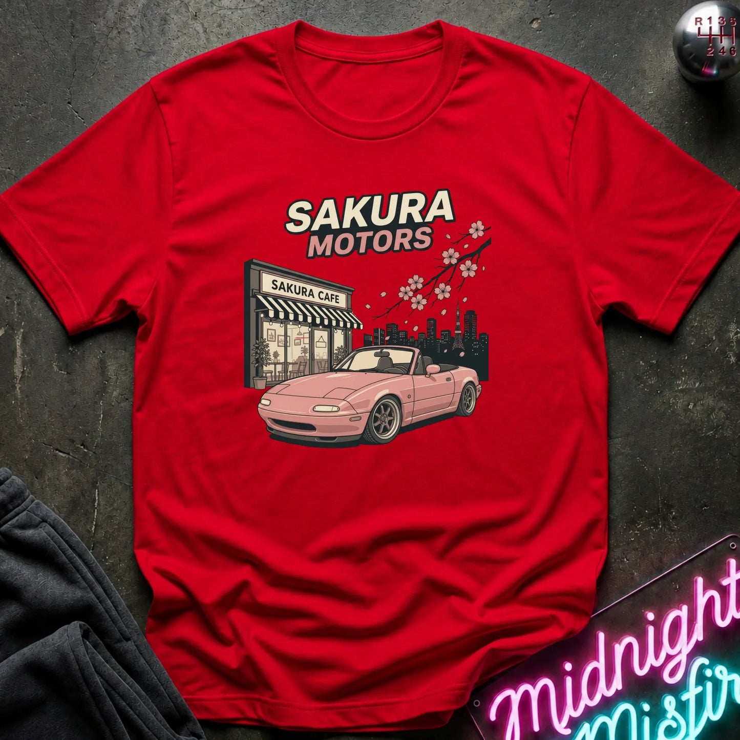 A red short-sleeve graphic T-shirt laid flat on a dark textured surface. The shirt features a printed design of a pink convertible car in front of a 'Sakura Motors' shop, with cherry blossoms and a city skyline.