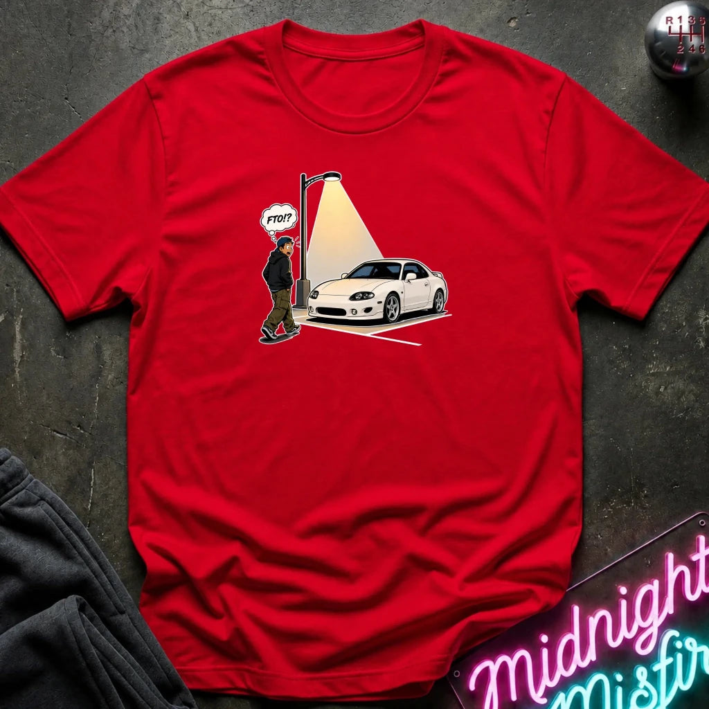A red short-sleeve crewneck t-shirt laid flat on a dark textured surface. The center of the shirt features a graphic print of a white sports car parked under a streetlamp with a spotlight effect, alongside a cartoon figure and a speech bubble 