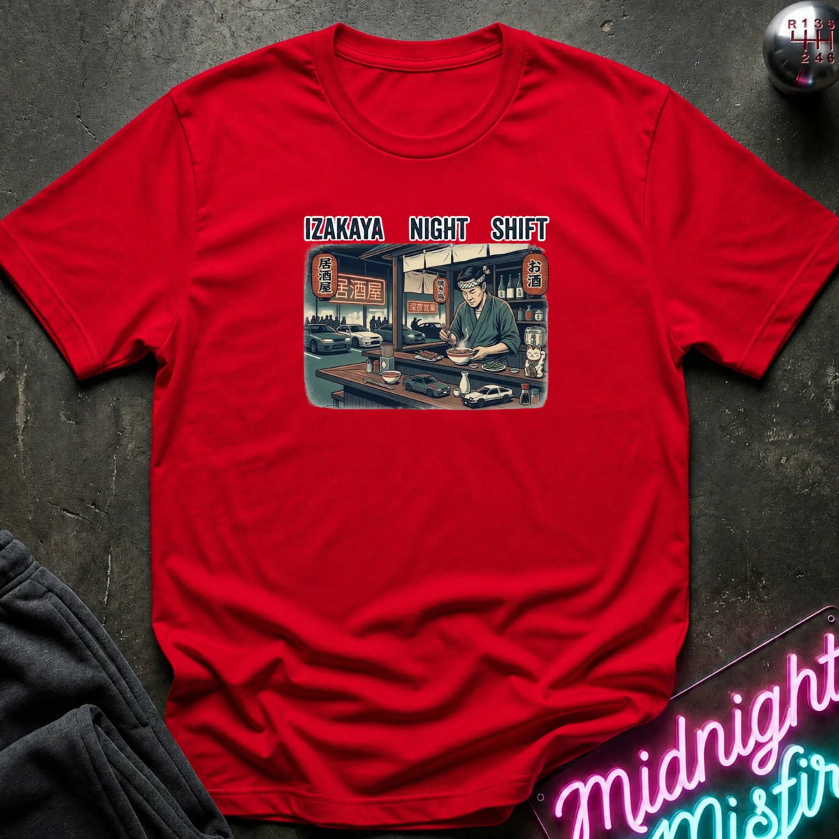 A red short-sleeve graphic t-shirt laid flat on a dark textured surface. The graphic features the text 'IZAKAYA NIGHT SHIFT' above an illustration of a man working late in a restaurant.