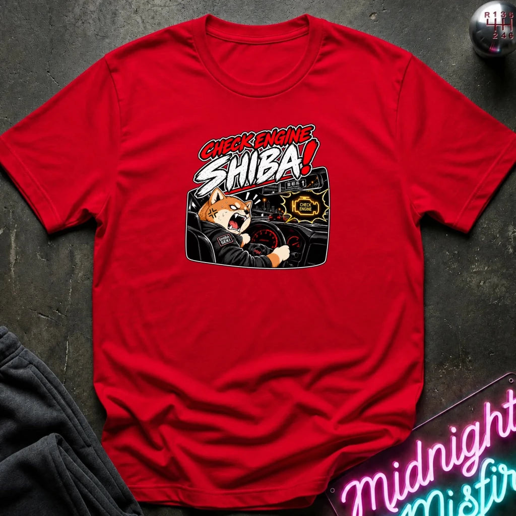 A red short-sleeve graphic T-shirt laid flat on a dark textured surface. The shirt features a cartoon Shiba Inu driving a car with the text 'CHECK ENGINE SHIBA!' in red, white, and black.