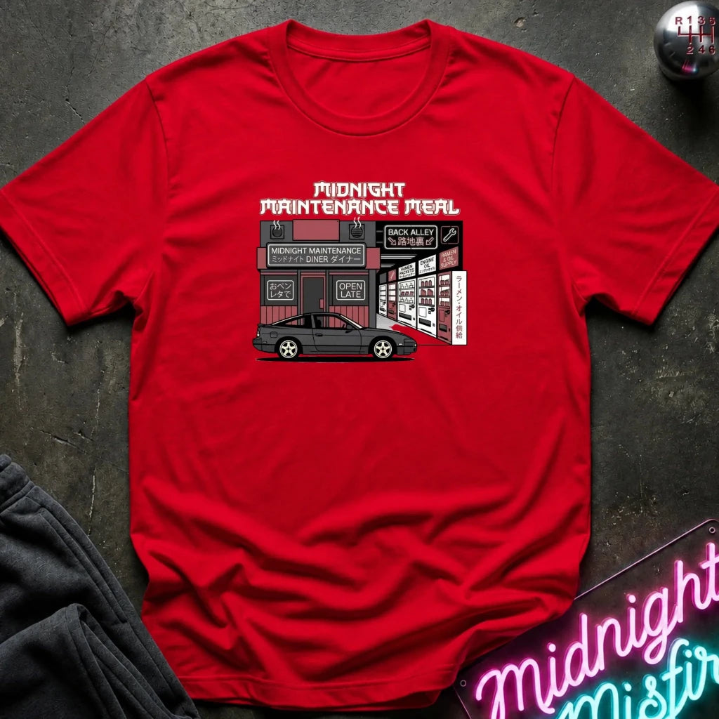 A red short-sleeve crewneck t-shirt featuring a graphic print of a garage scene with the text 'MIDNIGHT MAINTENANCE MEAL' and a black car illustration, laid flat on a dark textured surface with a neon sign and dark fabric partially visible.