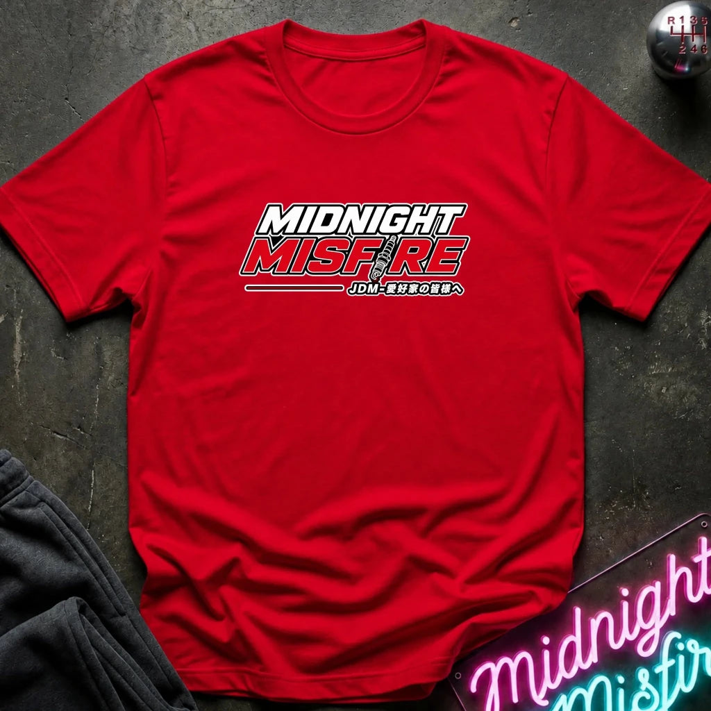 A red short-sleeve crewneck t-shirt with a 'MIDNIGHT MISFIRE' graphic printed on the chest, styled flat on a dark textured surface with a neon sign and dark pants in the background.