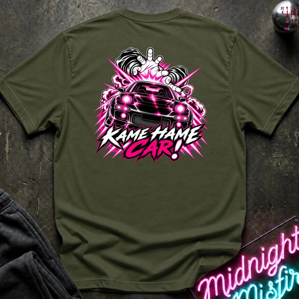 An olive green t-shirt laid flat on a dark textured surface, featuring a large graphic on the back with a black car, pink and white smoke effects, and the text 'KAME HAME CAR!'.