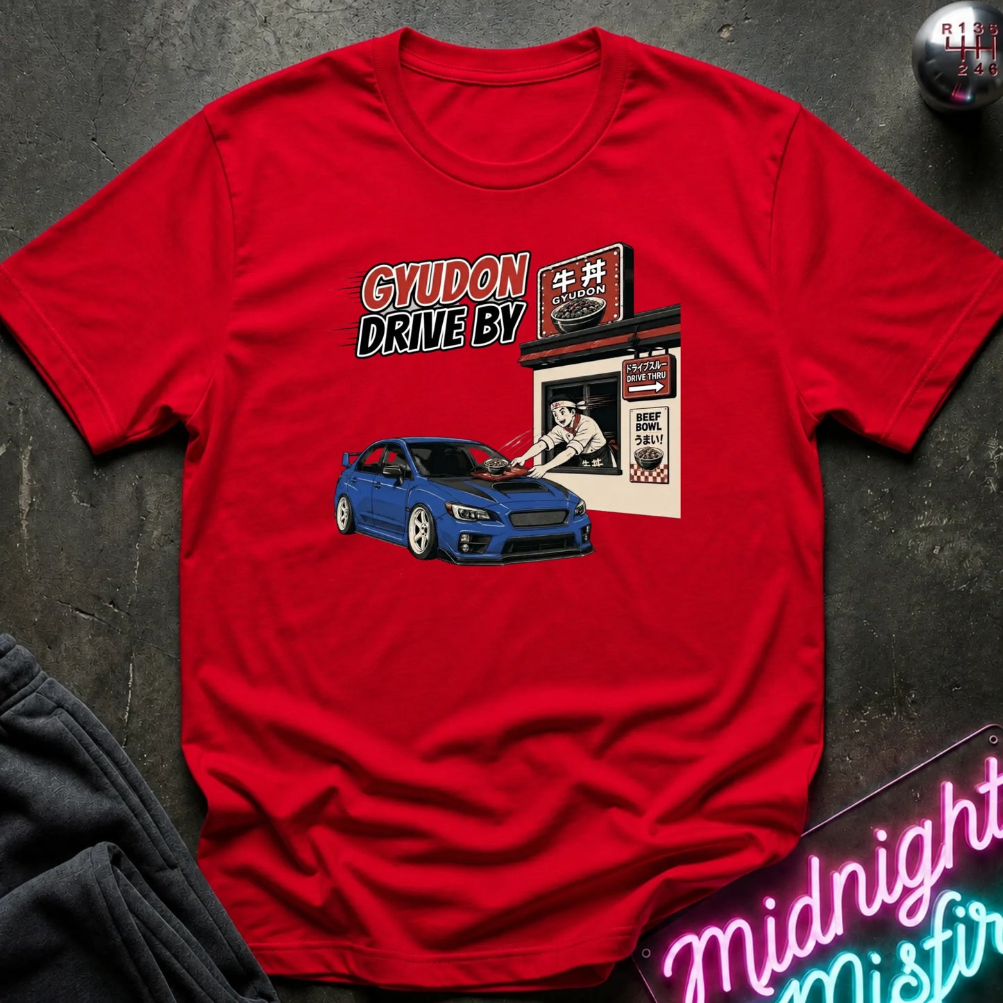 A red short-sleeve graphic T-shirt laid flat on a dark textured surface. The shirt features a printed design with the words 'GYUDON DRIVE BY' above a blue sports car and a small restaurant-style sign.