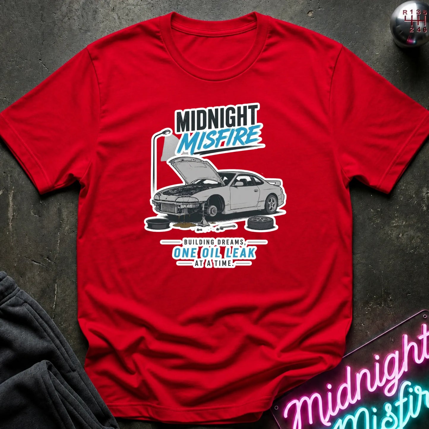 A red short-sleeve graphic T-shirt laid flat on a dark textured surface. The shirt features a printed design of a silver car with its hood open, accompanied by the text 'MIDNIGHT MISFIRE' and 'BUILDING DREAMS.