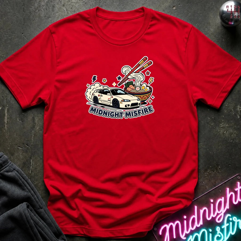 A red short-sleeve graphic T-shirt laid flat on a dark textured surface. The center graphic shows a white sports car with chopsticks, steam, and the text 'MIDNIGHT MISFIRE.