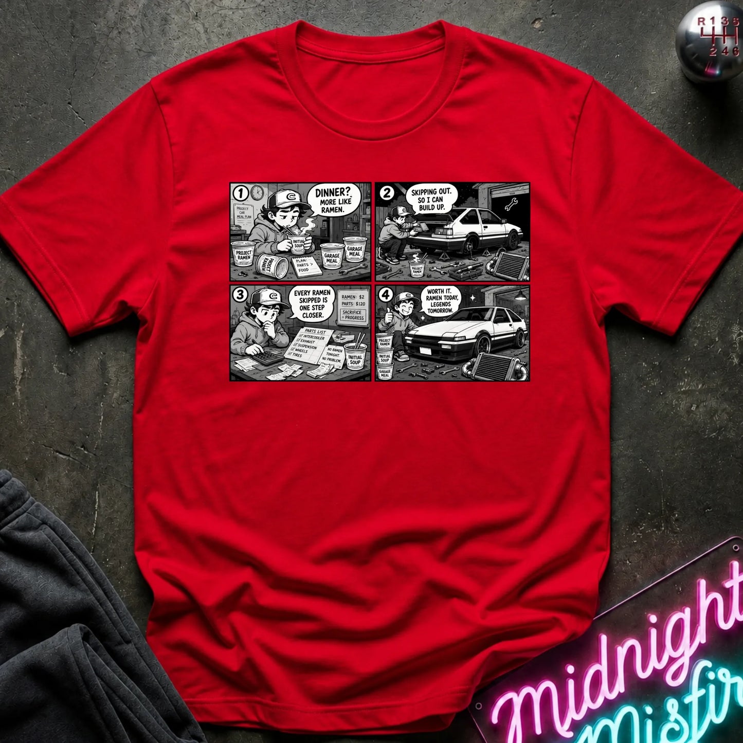 A red short-sleeve crewneck t-shirt featuring a black and white comic strip graphic on the chest, laid flat on a dark textured surface with a neon sign and dark pants partially visible.