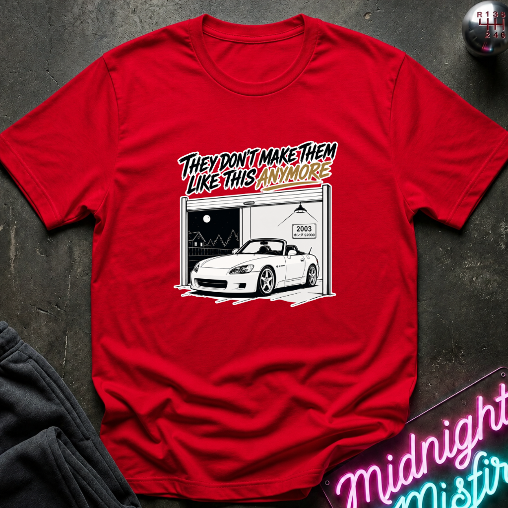 A red short-sleeve graphic T-shirt laid flat on a dark textured surface. The shirt features a white car garage scene with the text 'THEY DON'T MAKE THEM LIKE THIS ANYMORE' above a white sports car.