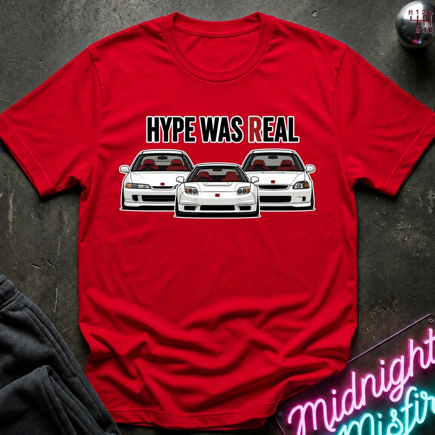 A red short-sleeve graphic t-shirt laid flat on a dark textured surface. The shirt features the text 'HYPE WAS REAL' above three white sports cars with red interiors.