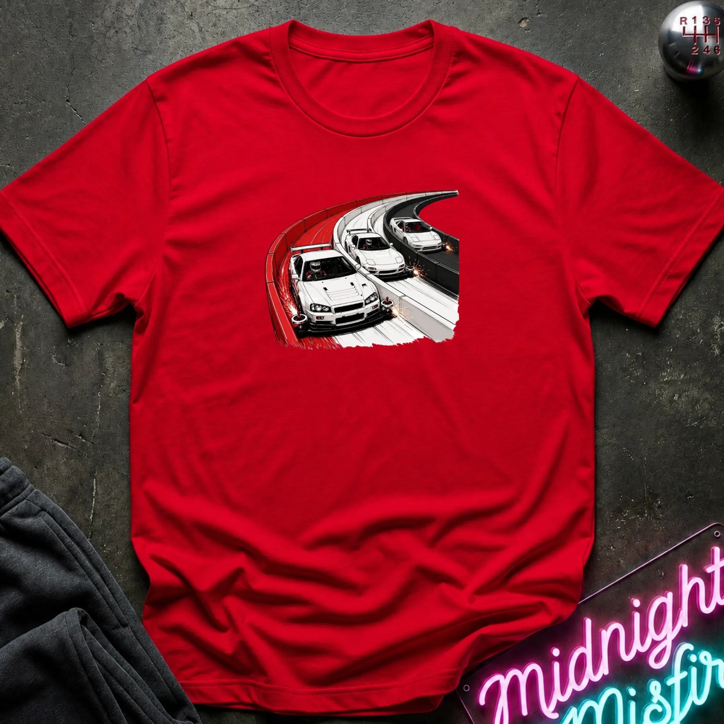 A bright red short-sleeve t-shirt laid flat on a dark textured surface, featuring a graphic print of three white sports cars racing on a curved track. 