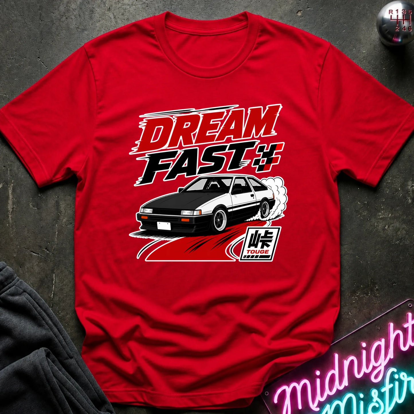A red short-sleeve graphic T-shirt laid flat on a dark textured surface. The shirt features a large front print with the words “DREAM FAST” in red and white, a black-and-white car, and a stylized “Fast” logo.