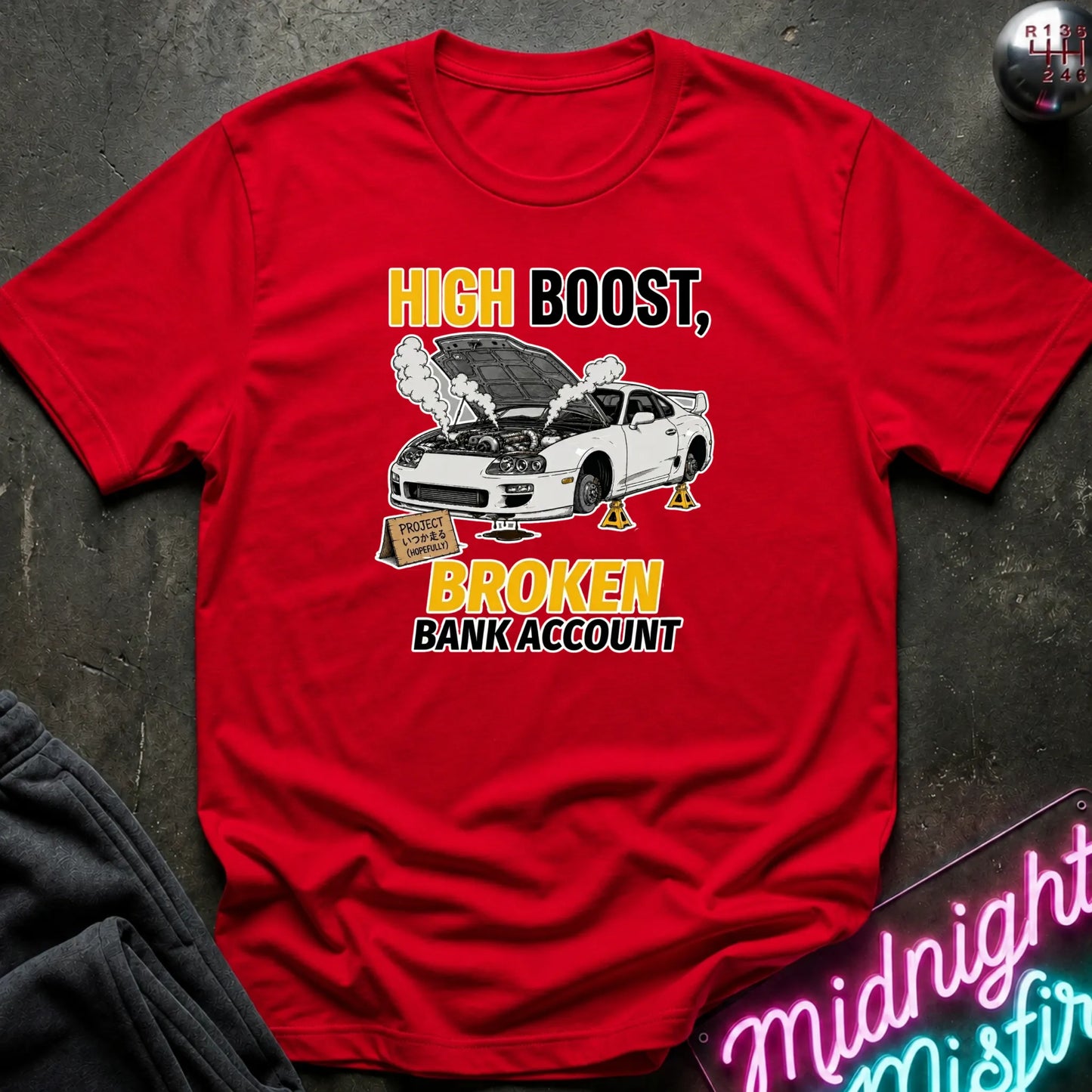 A red short-sleeve graphic t-shirt laid flat on a dark textured surface. The shirt features a humorous car graphic with the text 'HIGH BOOST, BROKEN BANK ACCOUNT' in yellow and black.