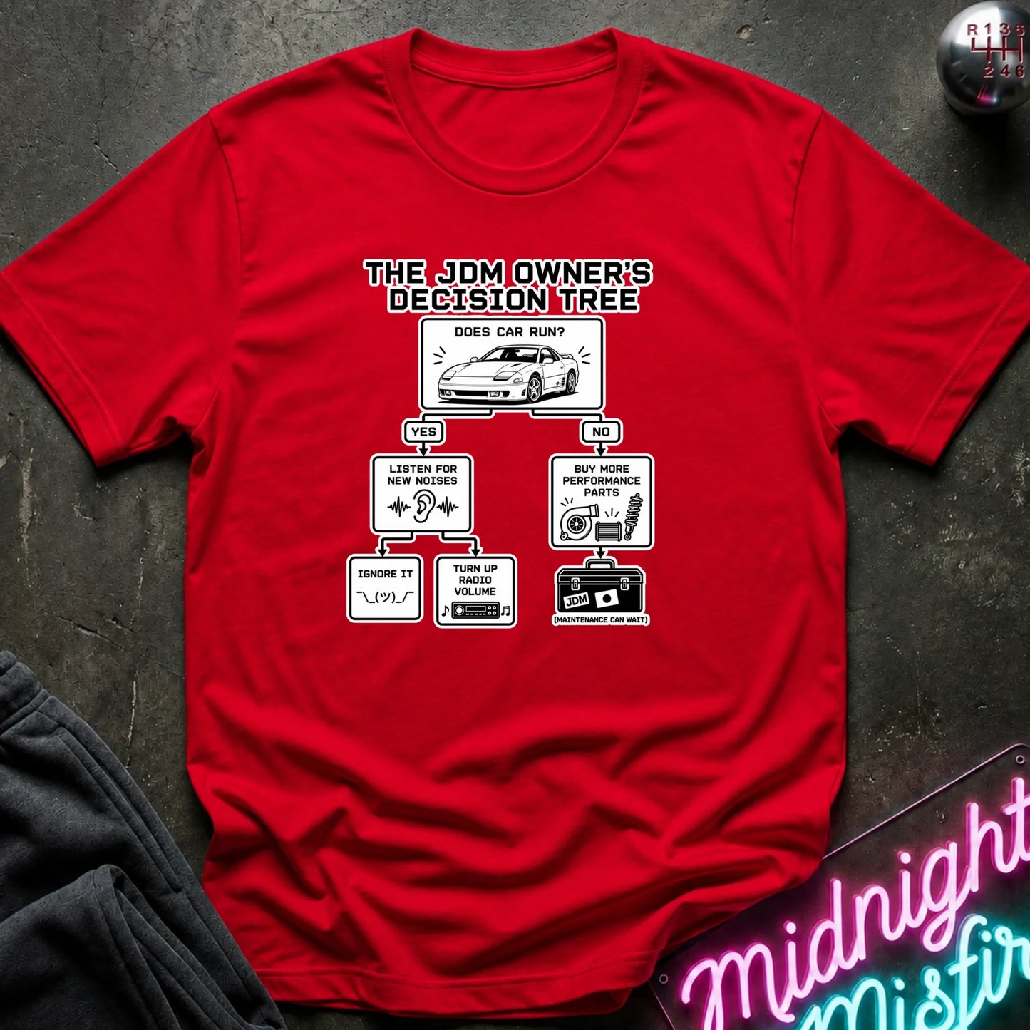 A red short-sleeve graphic T-shirt laid flat on a dark textured surface. The shirt features a white and black printed decision tree graphic titled 'THE JDM OWNER'S DECISION TREE' with illustrations and text related to car care and maintenance.
