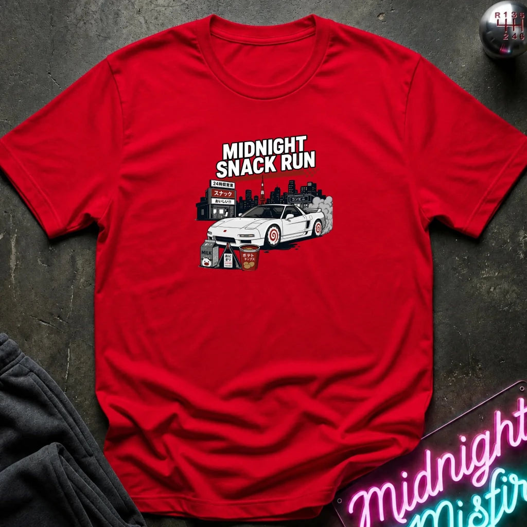 A red short-sleeve crewneck t-shirt with a graphic print of a white sports car and the text 'MIDNIGHT SNACK RUN' on the chest, styled flat on a dark textured background with a neon sign and dark sweatpants partially visible.