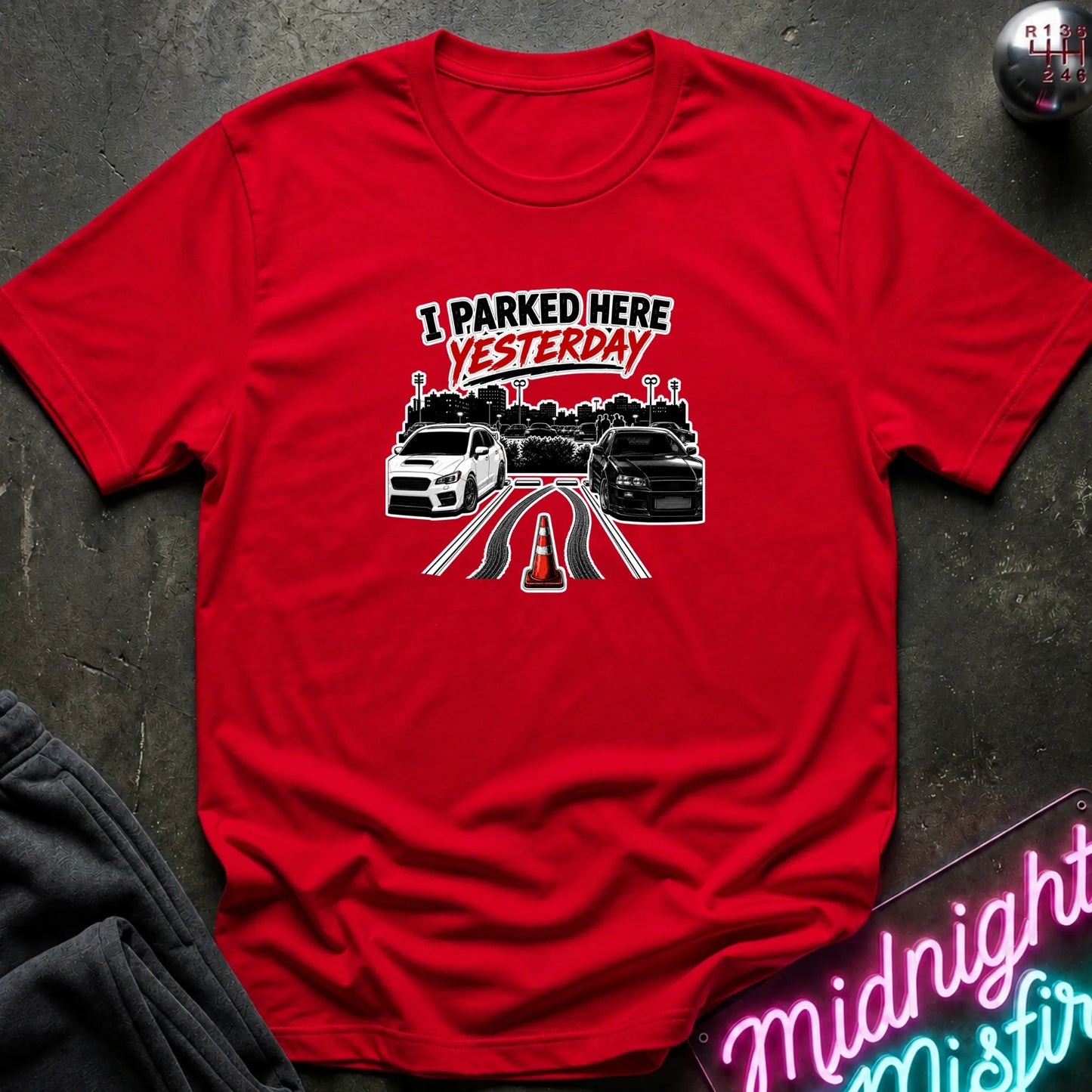 A red short-sleeved t-shirt  that has 2 cars parked in a space in between empty space with skid marks.
