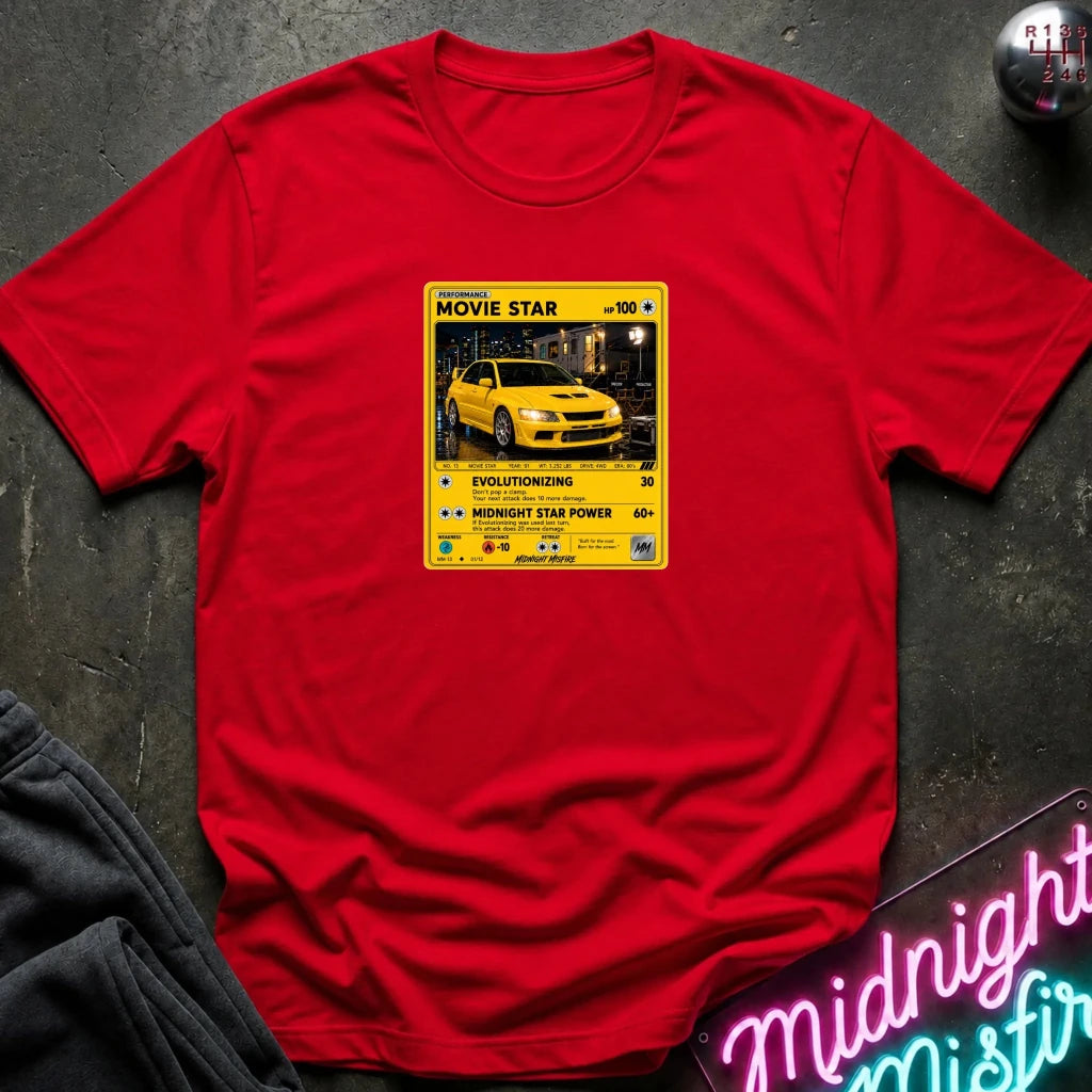 red short-sleeve t-shirt laid flat on a dark textured surface, featuring a yellow and black retro-style graphic on the chest with a yellow car photo and text including 'MOVIE STAR'. 