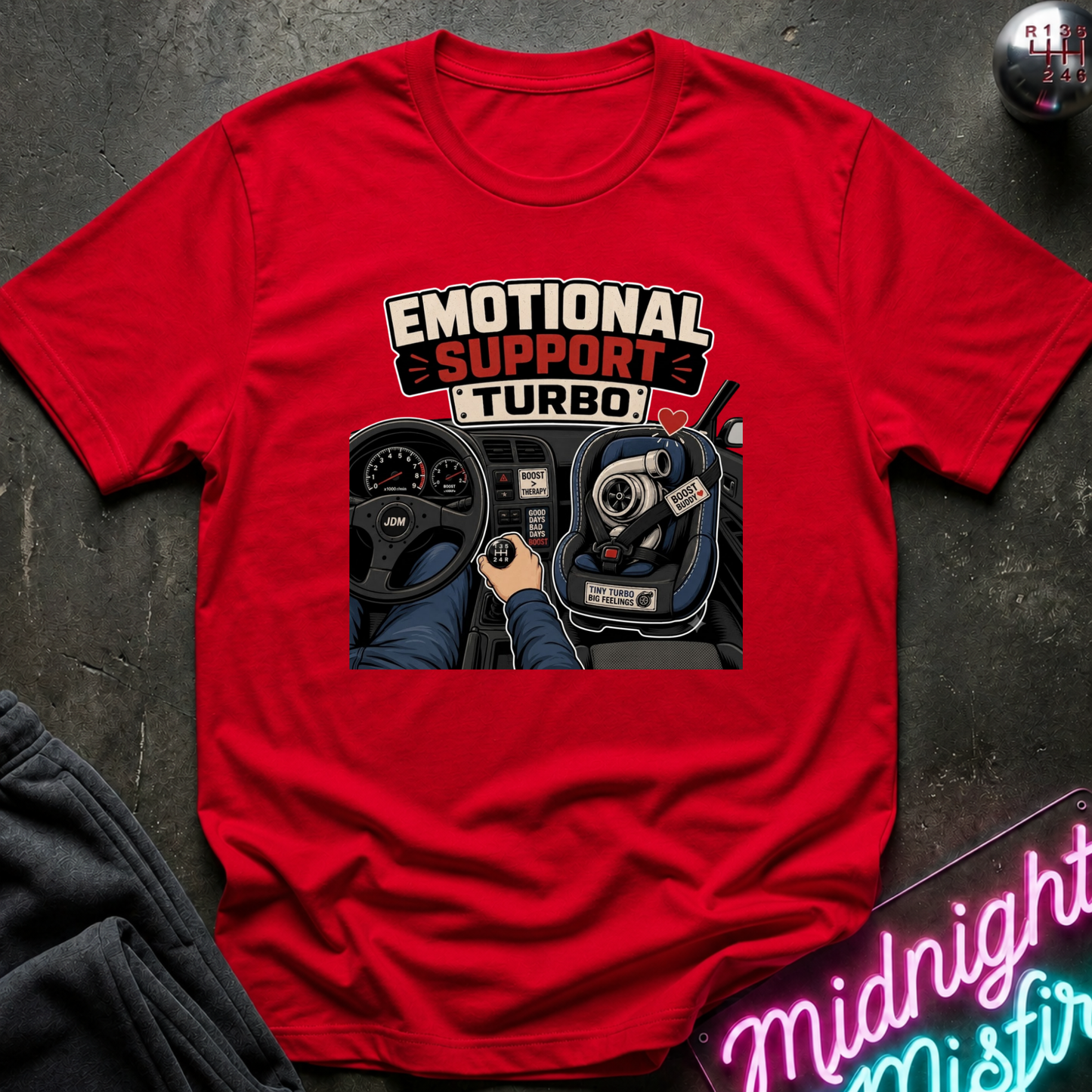 A red short-sleeve graphic T-shirt laid flat on a dark textured surface. The shirt features a large front print with the words 'EMOTIONAL SUPPORT TURBO' above an illustration of a hand gripping a steering wheel in a car interior.