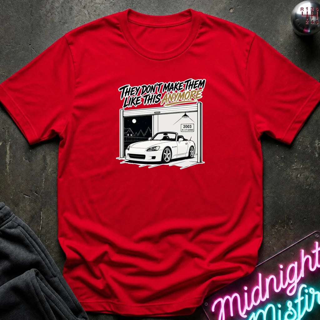 A red short-sleeve graphic t-shirt laid flat on a dark textured surface. The graphic features a white sports car inside a garage with the text 'THEY DON'T MAKE THEM LIKE THIS ANYMORE' in a stylized black, white, and yellow font.