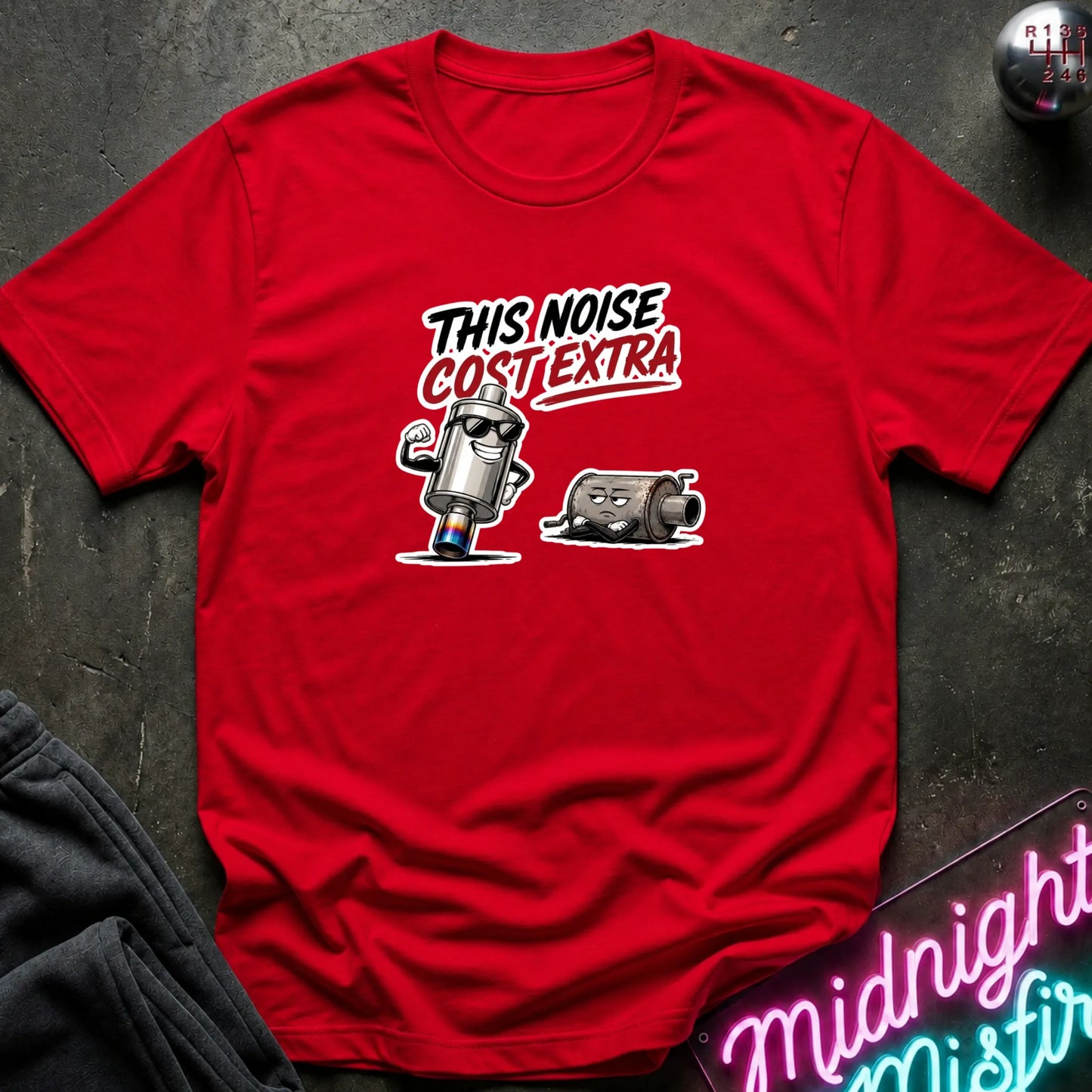 A red short-sleeve graphic T-shirt laid flat on a dark textured surface. The shirt features a text 'THIS NOISE COST EXTRA' in white, black, and red.