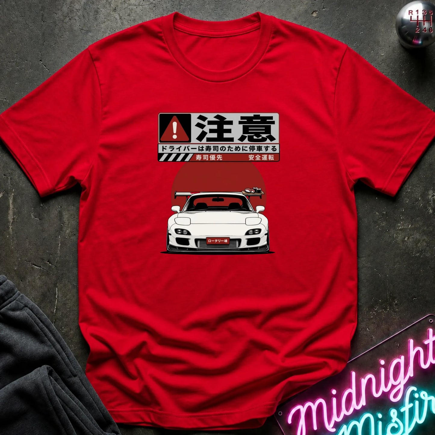 A red short-sleeve crewneck t-shirt laid flat on a dark textured surface. The shirt features a graphic print of a white sports car under a Japanese warning sign design.