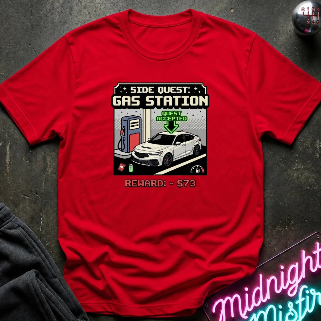 A red short-sleeve graphic t-shirt laid flat on a dark textured surface. The graphic depicts a retro video game side quest for a gas station with a white car, pump, and text reading 'SIDE QUEST: GAS STATION' and 'REWARD: $73'.